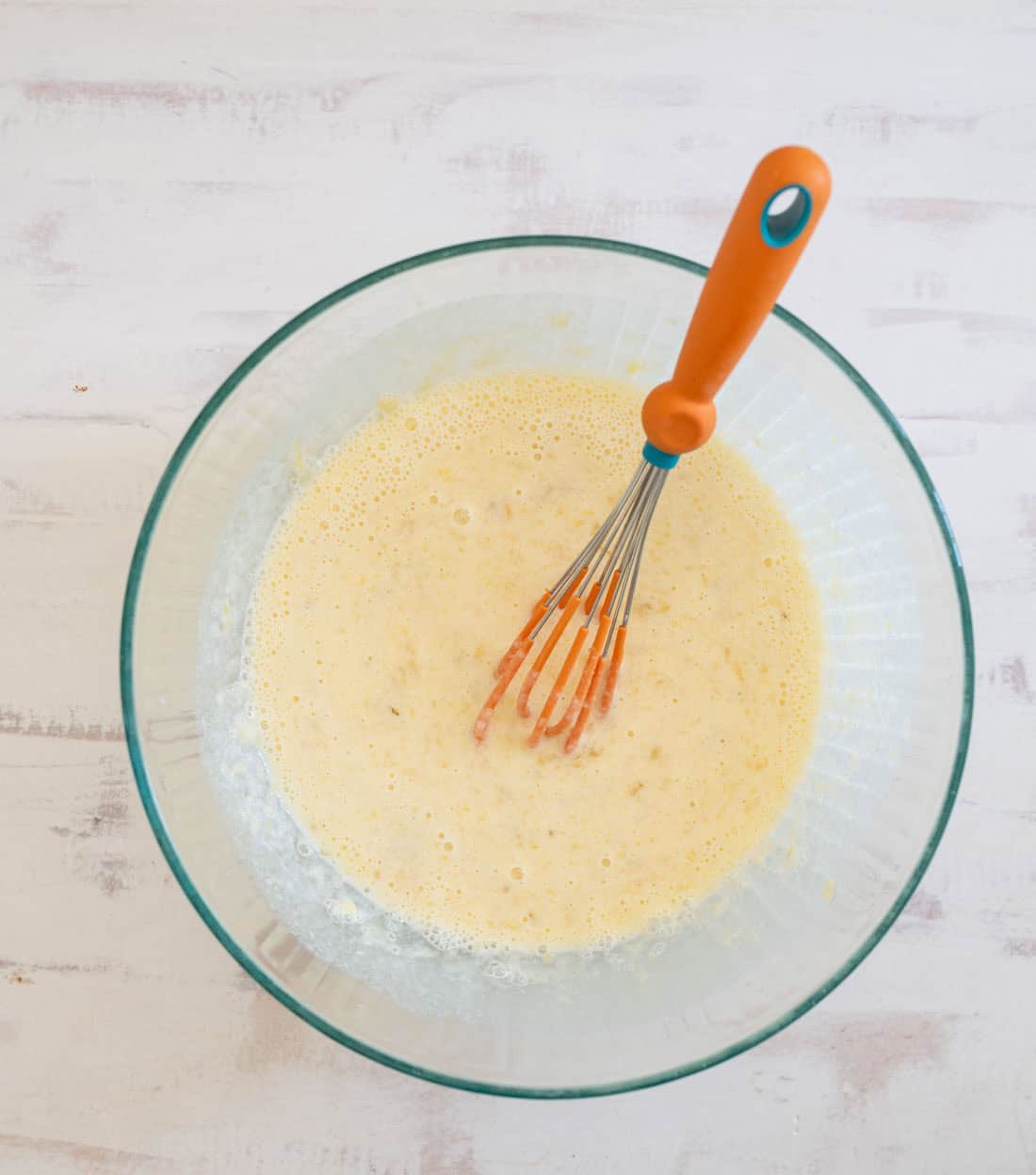 A glass bowl filled with a light yellow batter is shown on a white surface. An orange-handled whisk rests inside the bowl, partially submerged in the mixture. The batter appears smooth and slightly frothy.