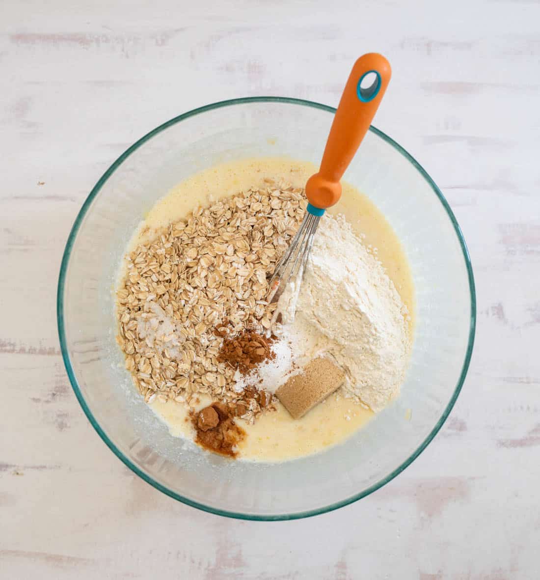 Glass bowl with batter, oats, flour, brown sugar, cinnamon, baking powder, and salt. Orange-handled whisk stands upright. All on a white surface.