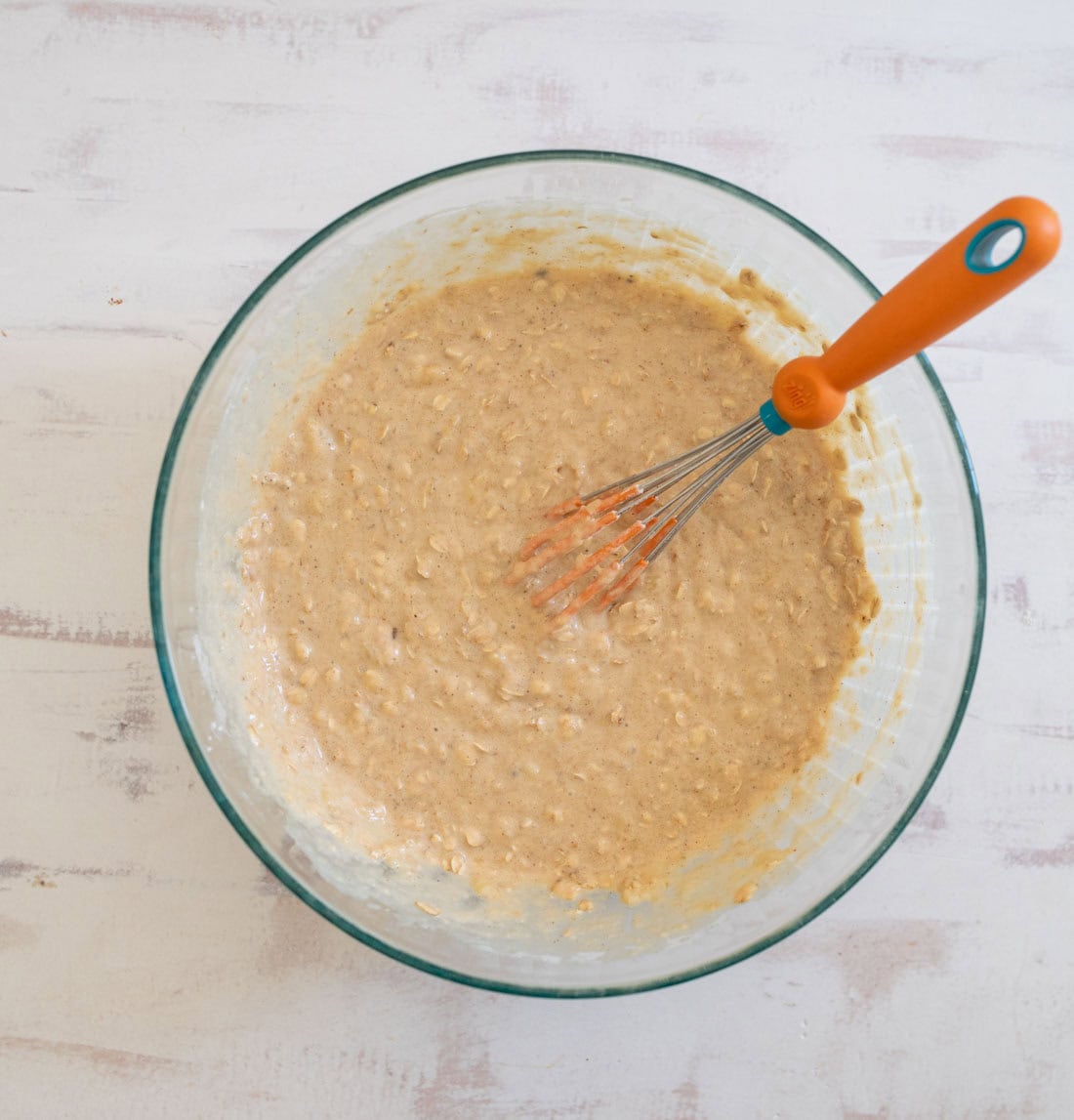 A glass bowl filled with a thick, beige batter on a white countertop. A bright orange whisk is resting inside the bowl, partially submerged in the mixture. The surface has a rustic, slightly weathered appearance.
