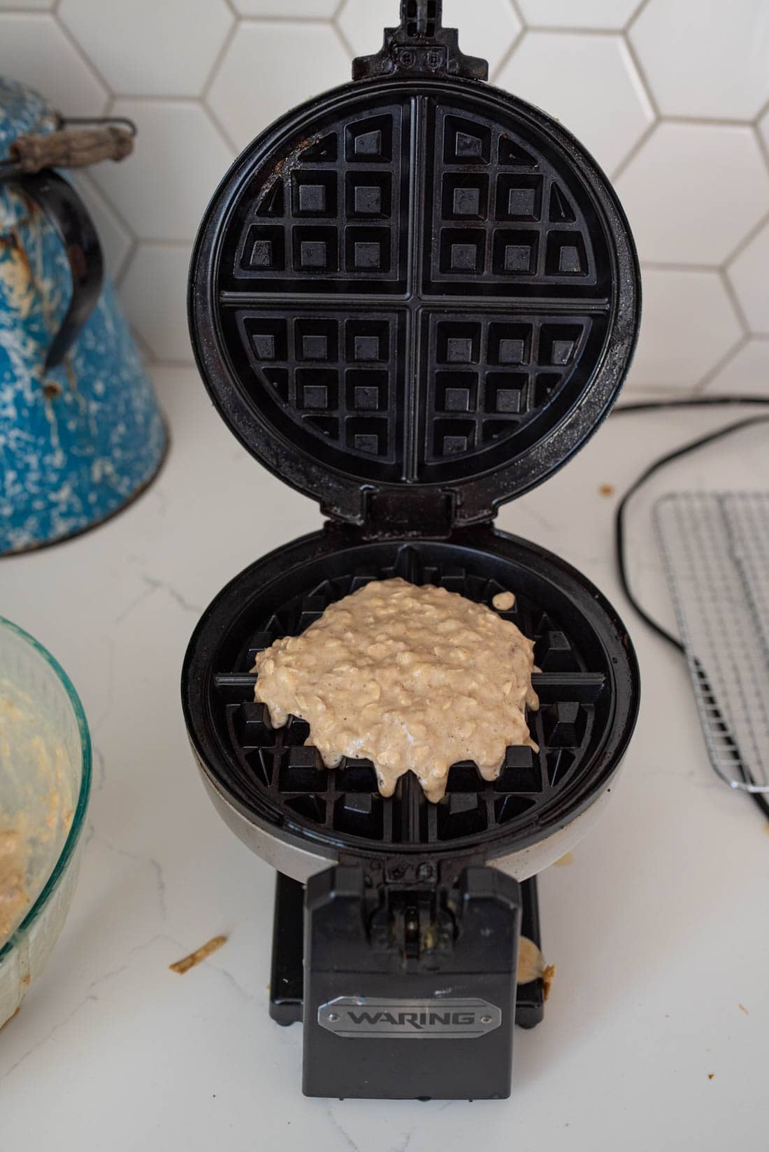 A waffle maker with uncooked batter is open on a kitchen counter. The appliance is black, and the counter has a white hexagon-tiled backsplash. A glass mixing bowl and a blue rustic kettle are nearby.
