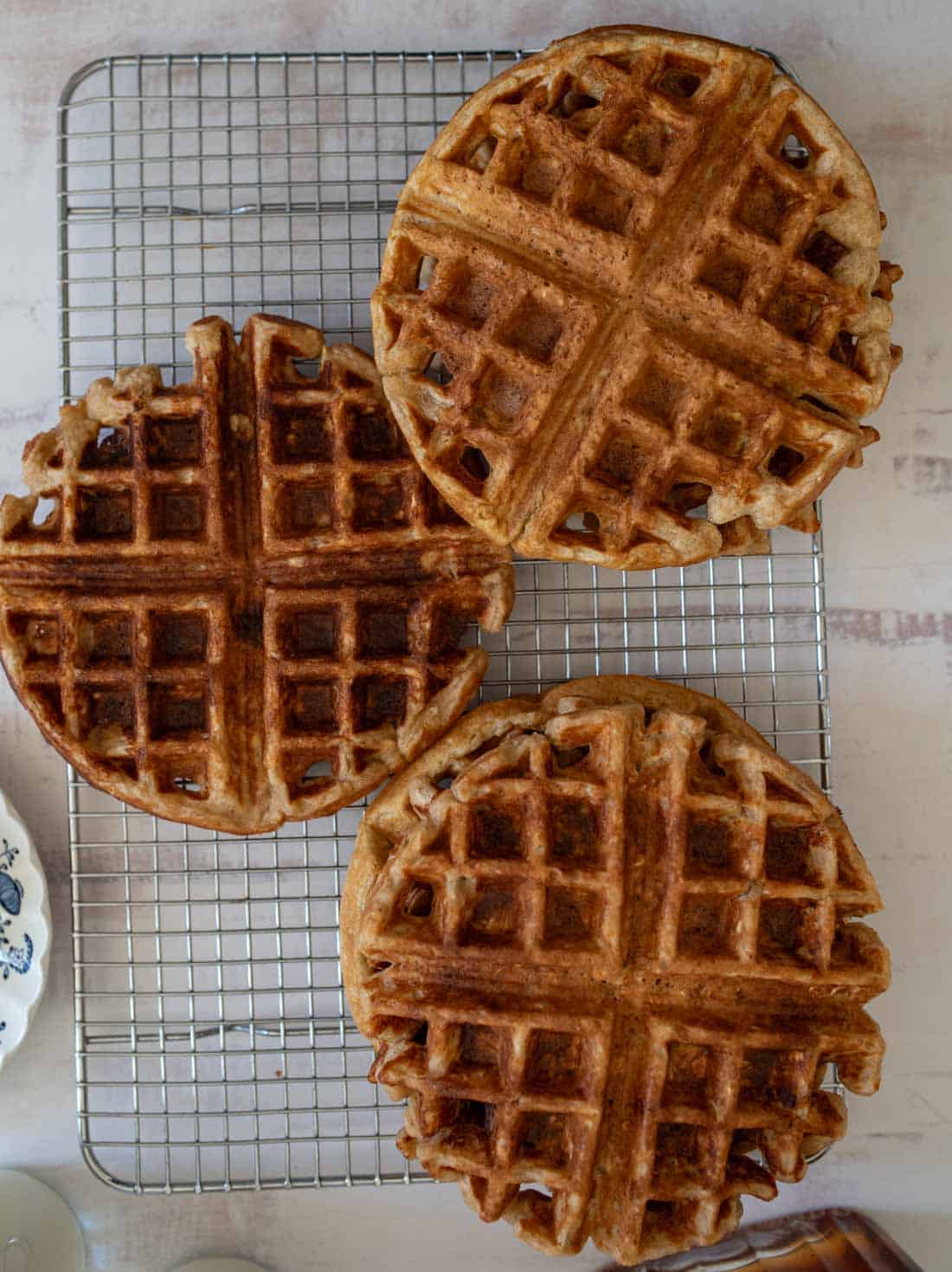 Three golden-brown waffles are cooling on a wire rack. The waffles have a classic grid pattern and are placed on a light-colored surface. Nearby, a portion of a decorative plate is visible.