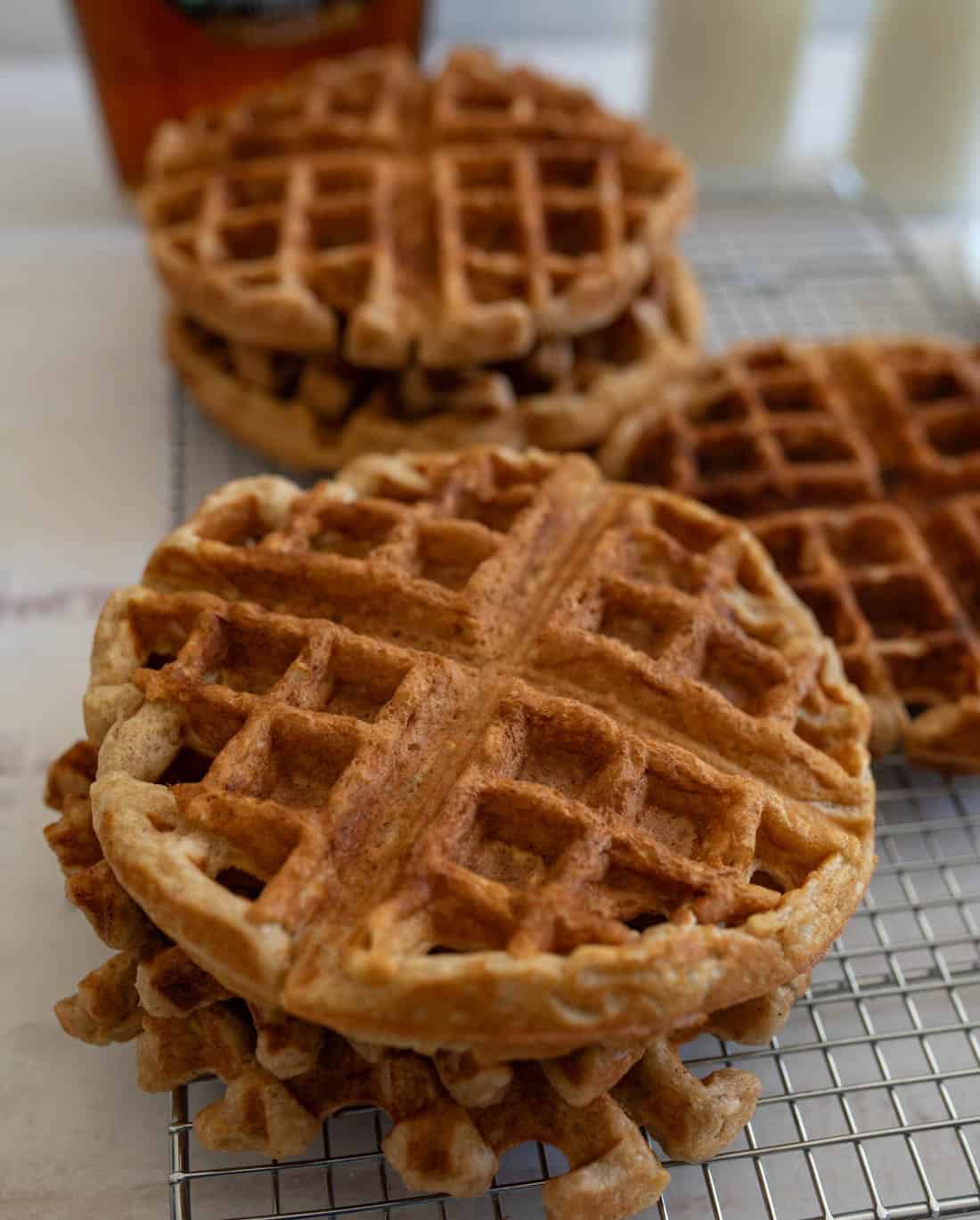 Golden-brown waffles stacked on a cooling rack, with a blurred background including a jar and a bottle of milk.