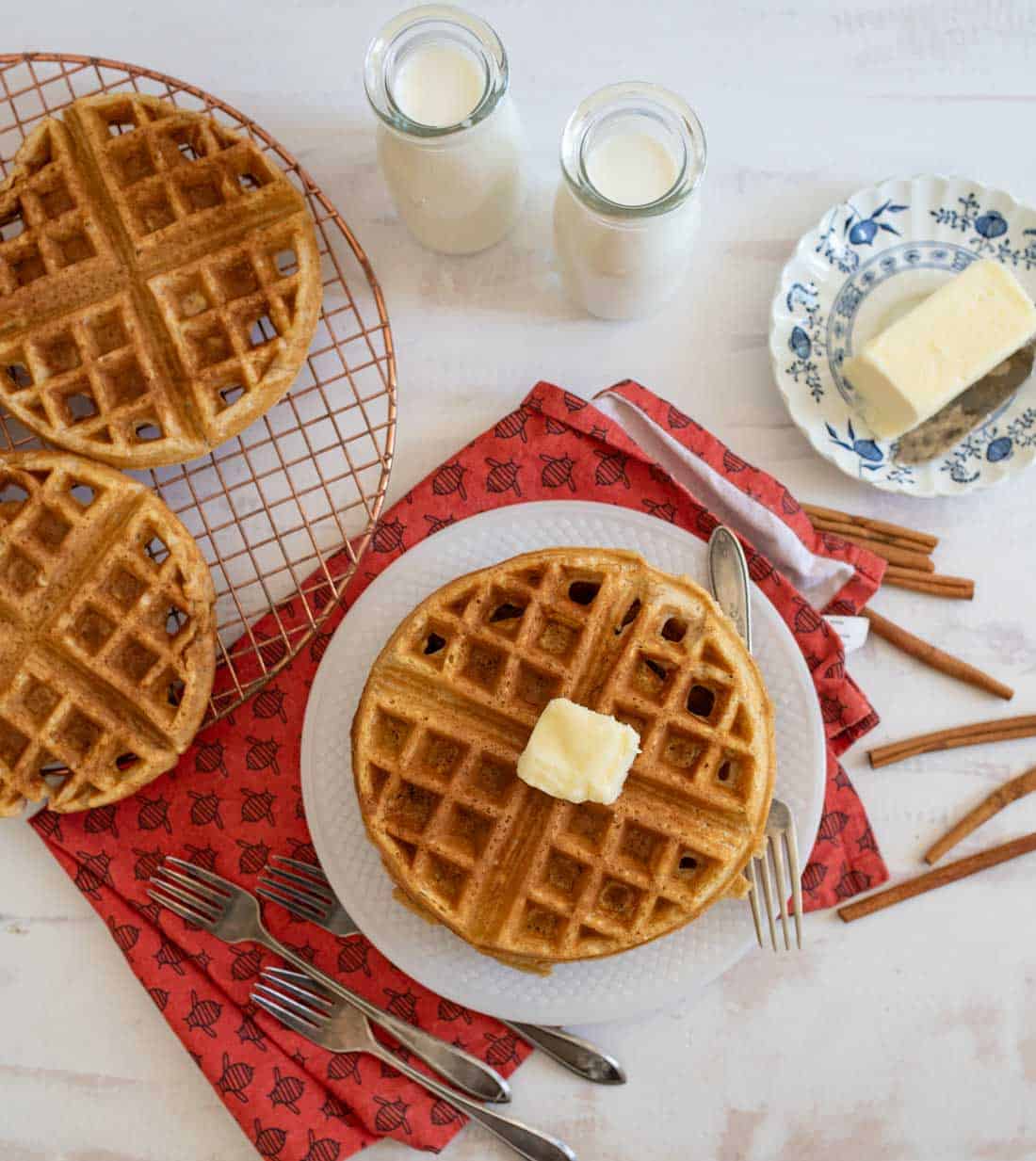 A breakfast setting with waffles on a white plate, topped with a pat of butter. Two glass bottles of milk, a piece of butter on a floral plate, cinnamon sticks, and a red napkin with forks surround the scene.