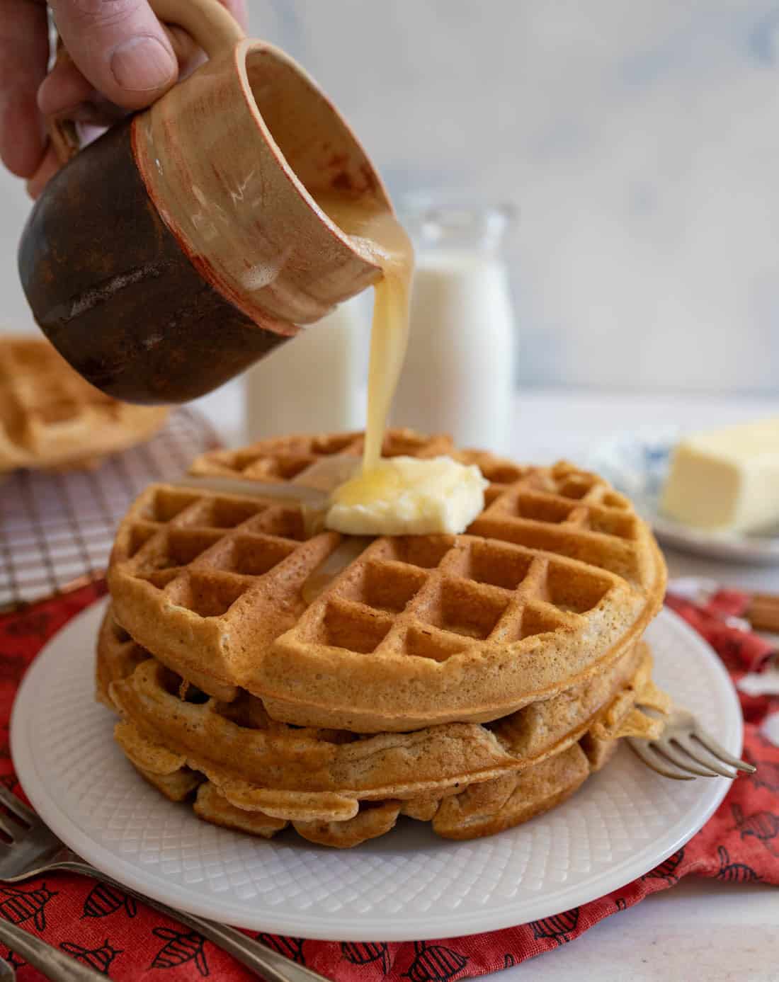 A hand pours syrup from a rustic cup onto a stack of waffles topped with butter on a white plate. A red cloth and a bottle of milk are in the background, along with a pat of butter and a fork.