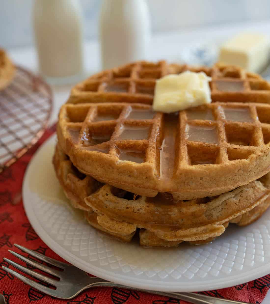 A stack of cinnamon waffles topped with a pat of butter sits on a white plate with syrup drizzled over them. In the background, there are two milk bottles, a block of butter, a fork, and a red napkin.