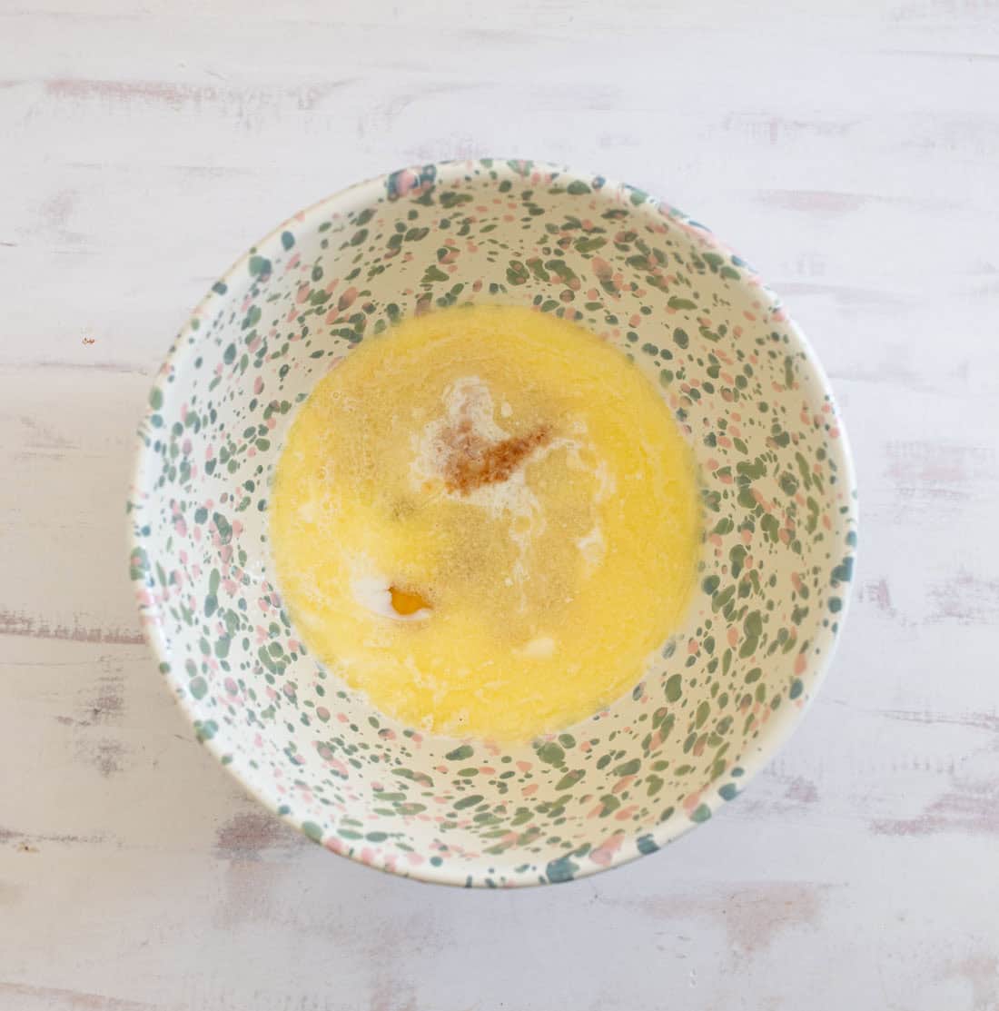 A speckled bowl containing a mix of melted butter, sugar, and a hint of cinnamon, set on a light wood surface.