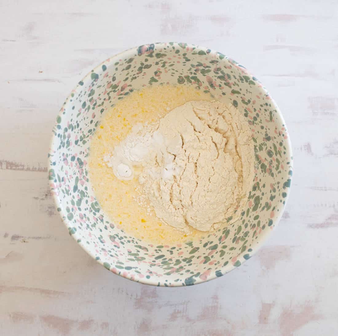 A patterned bowl on a white surface contains flour and a yellow liquid mixture, likely batter ingredients, yet to be mixed.