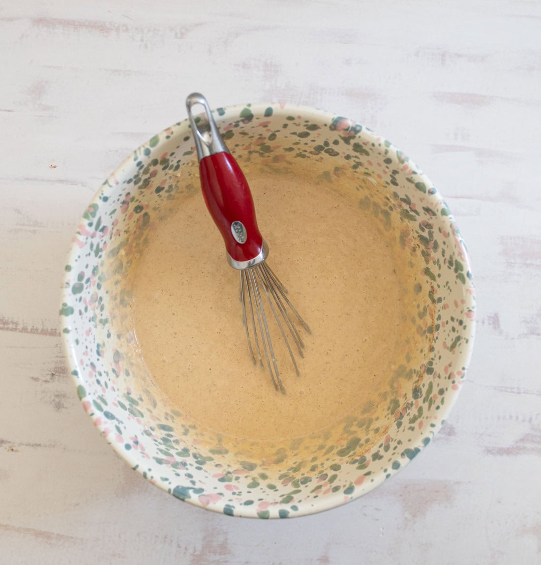 A colorful mixing bowl with a floral pattern contains a light brown batter. A metal whisk with a red handle rests inside the bowl on a light wooden surface.