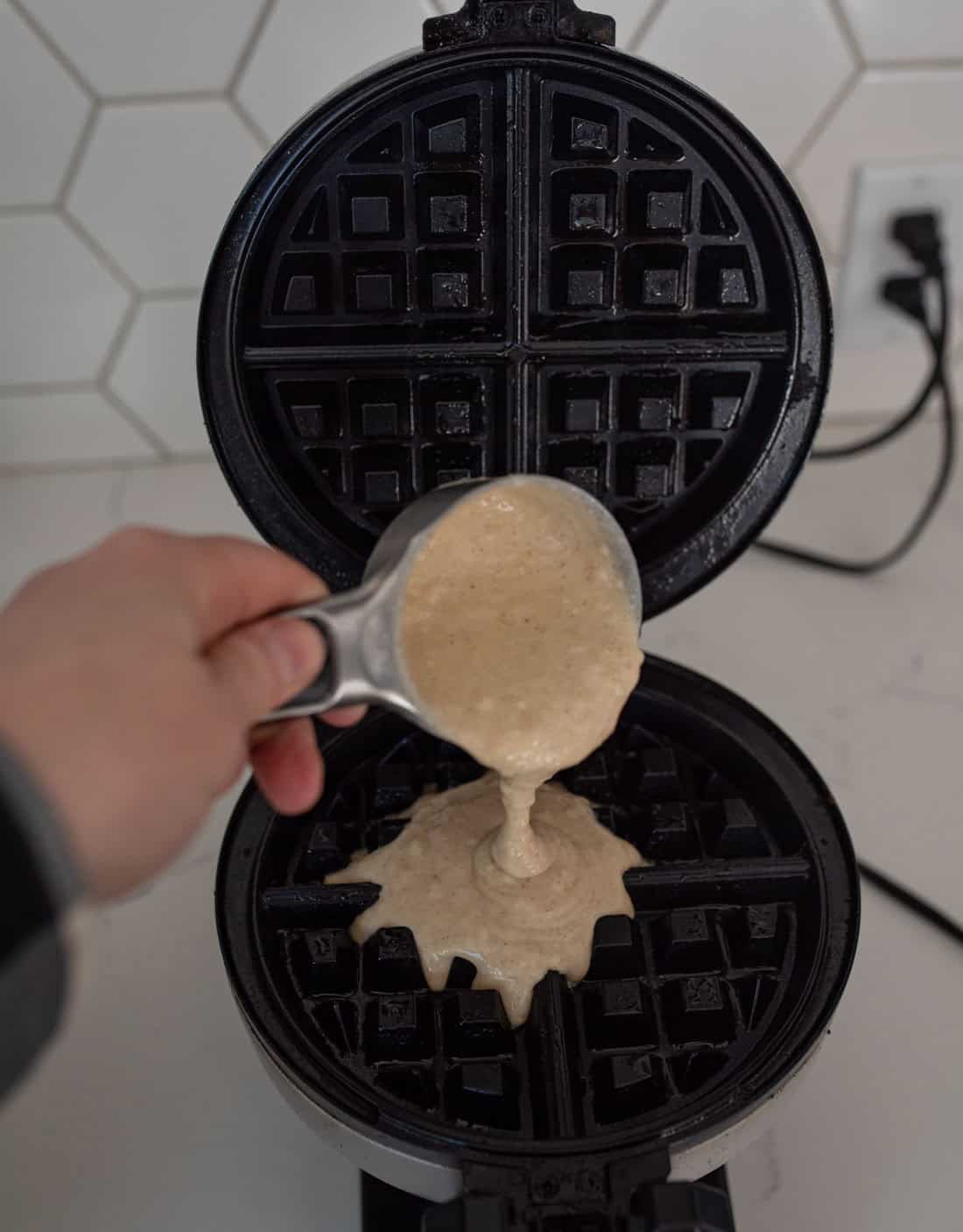 Person pouring waffle batter from a measuring cup onto a waffle iron. The open waffle iron is on a white countertop with a hexagonal tile backsplash in the background. An electrical outlet is visible on the wall.
