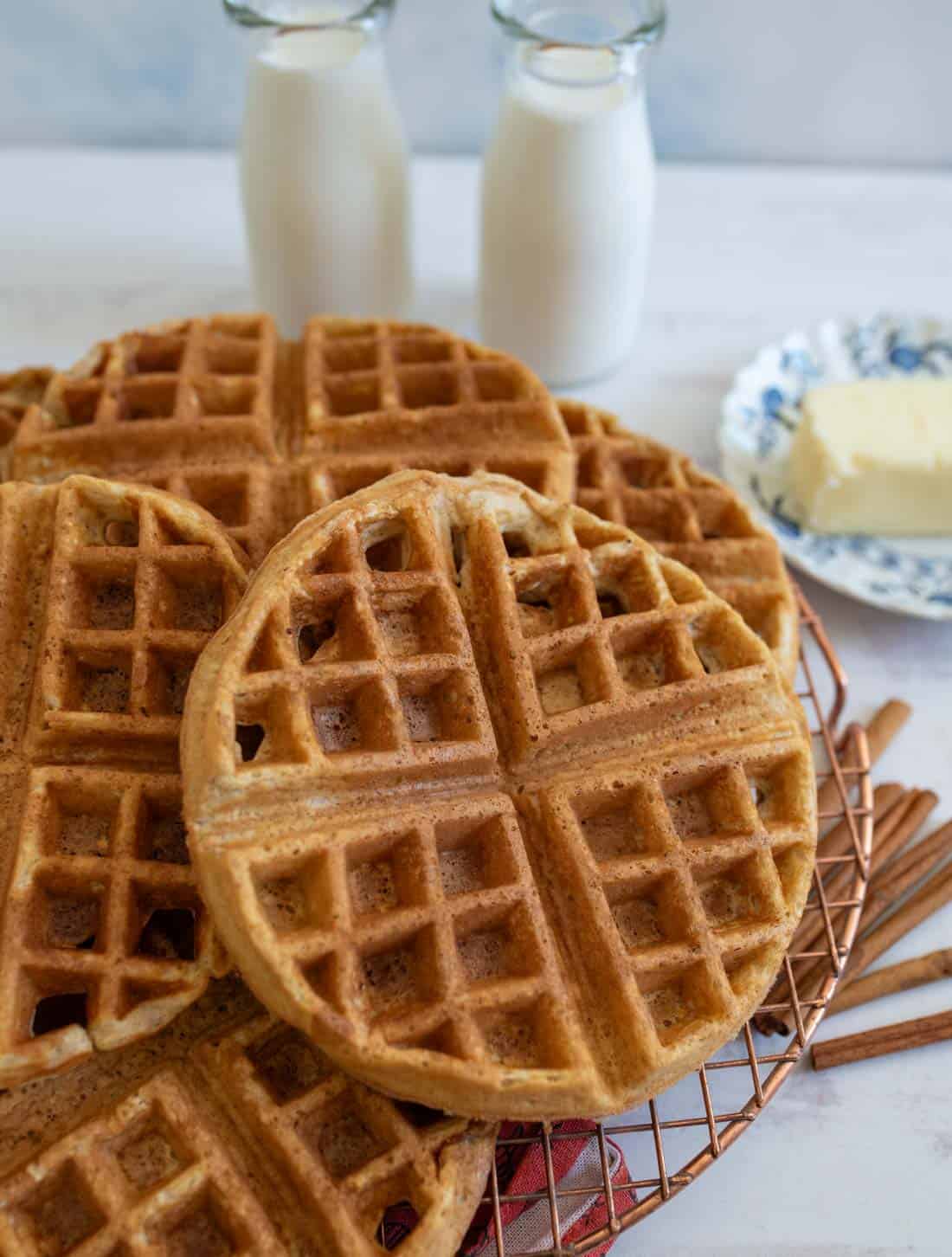 Waffles stacked on a wire rack, with two bottles of milk and a plate of butter in the background. Cinnamon sticks are scattered around, suggesting a cozy breakfast scene.