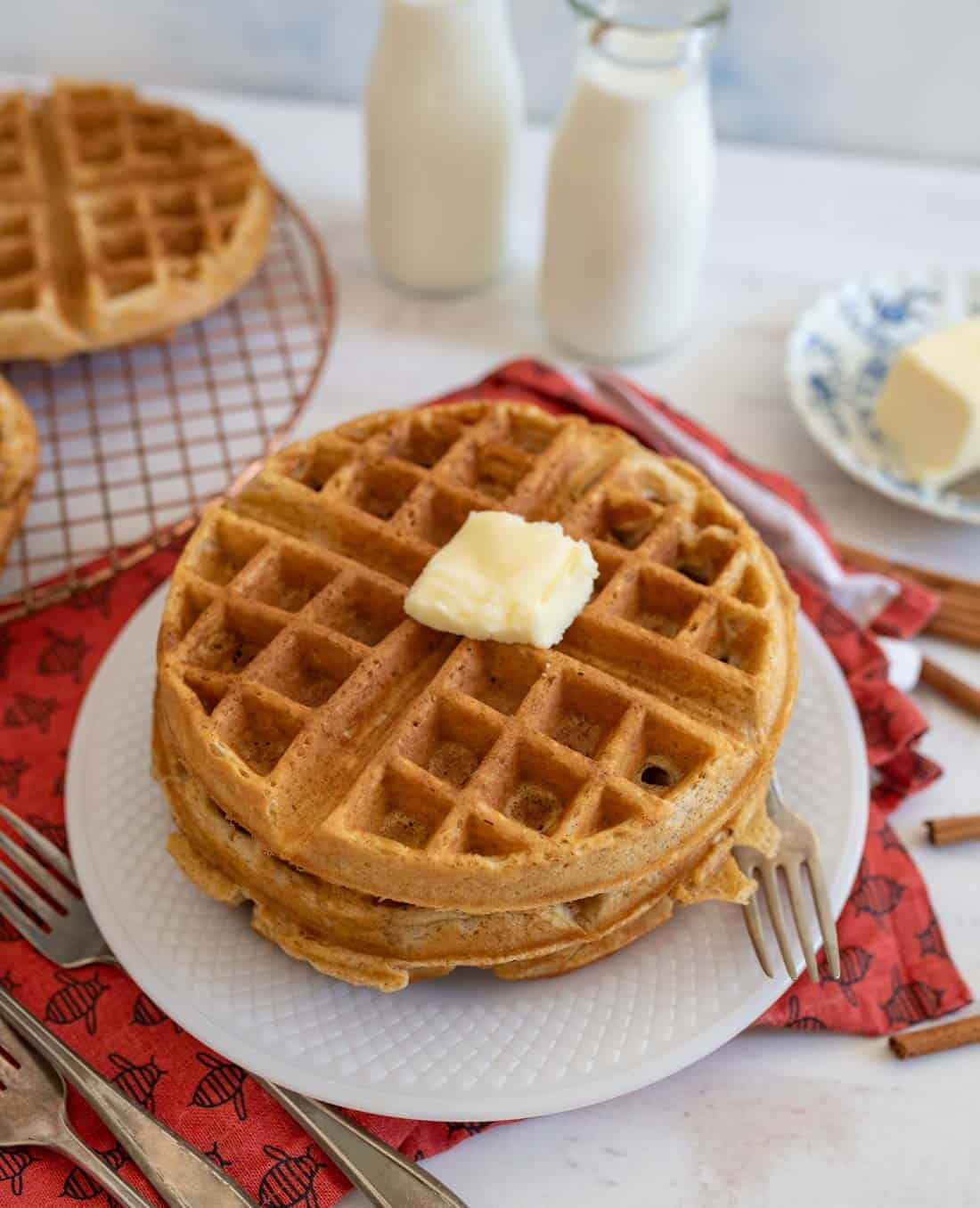 A stack of waffles topped with a pat of butter on a white plate. Two forks rest on the plates edge. In the background, a cooling rack with waffles, two bottles of milk, and a plate with more butter are visible, all set on a red cloth.