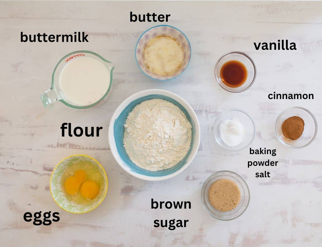 An assortment of baking ingredients is laid out on a white surface, including buttermilk, butter, vanilla, flour, eggs, brown sugar, cinnamon, and baking powder plus salt, each in separate bowls or measuring cups. Labels identify each ingredient.