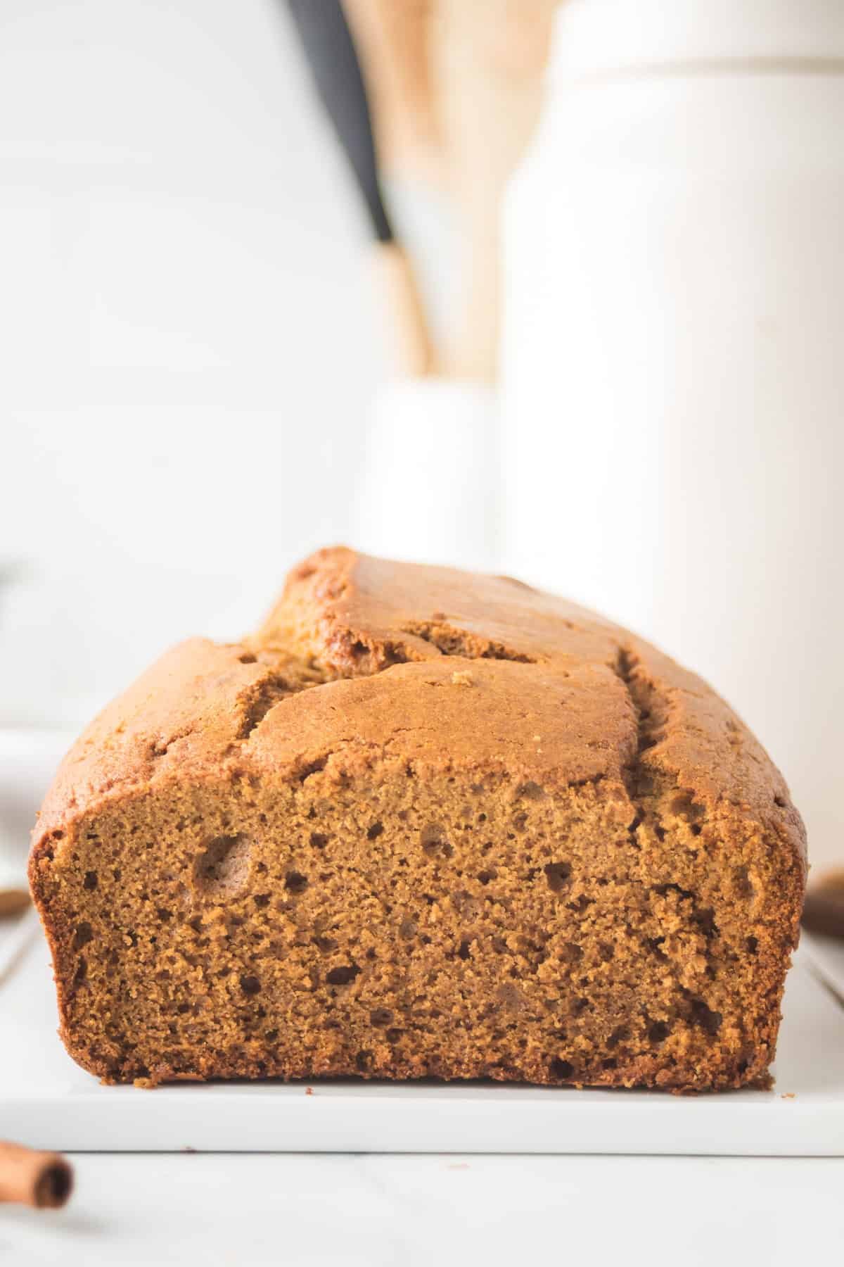 A close-up of a loaf of bread on a white surface, showcasing its golden-brown crust and fluffy texture. In the blurred background, there are light-colored jars and kitchen utensils.