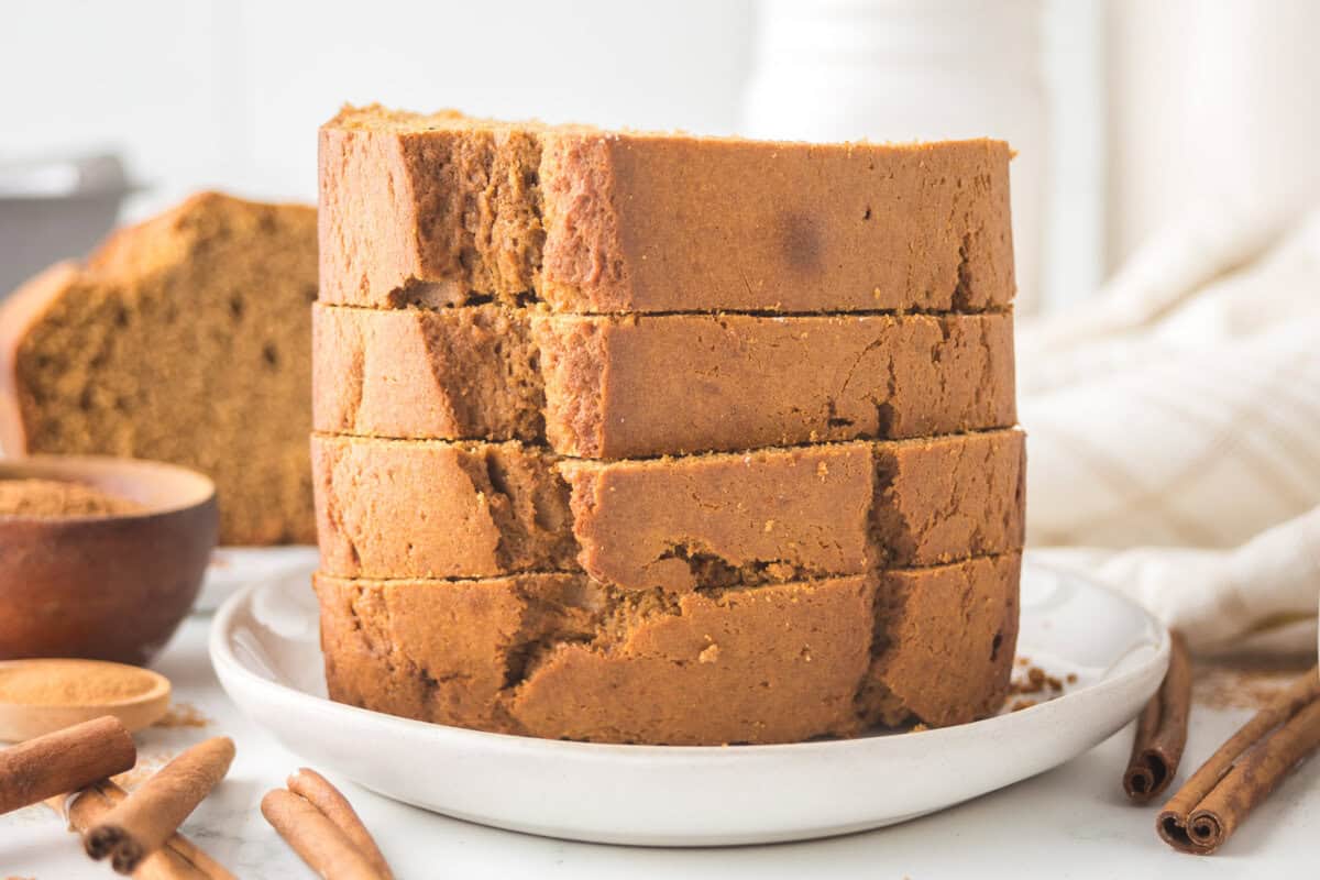 Sliced gingerbread loaf stacked on a white plate, surrounded by cinnamon sticks and a bowl of brown sugar. A loaf of bread and a white cloth are in the background. The setting is bright, conveying a cozy and inviting atmosphere.