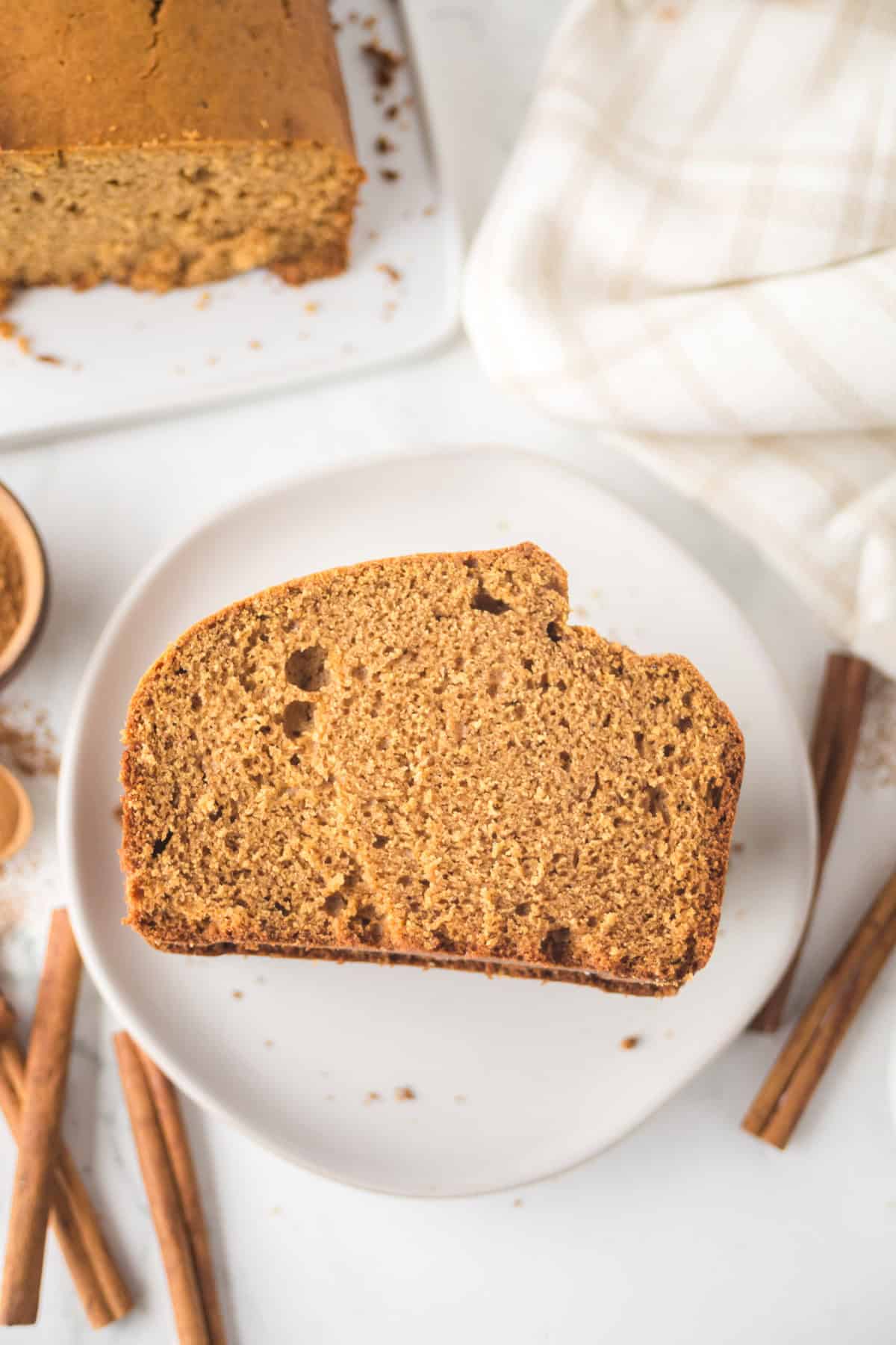 A slice of brown gingerbread loaf on a white plate, surrounded by cinnamon sticks and a light-checkered cloth. More bread is visible in the background. The setting suggests a cozy, homemade treat.