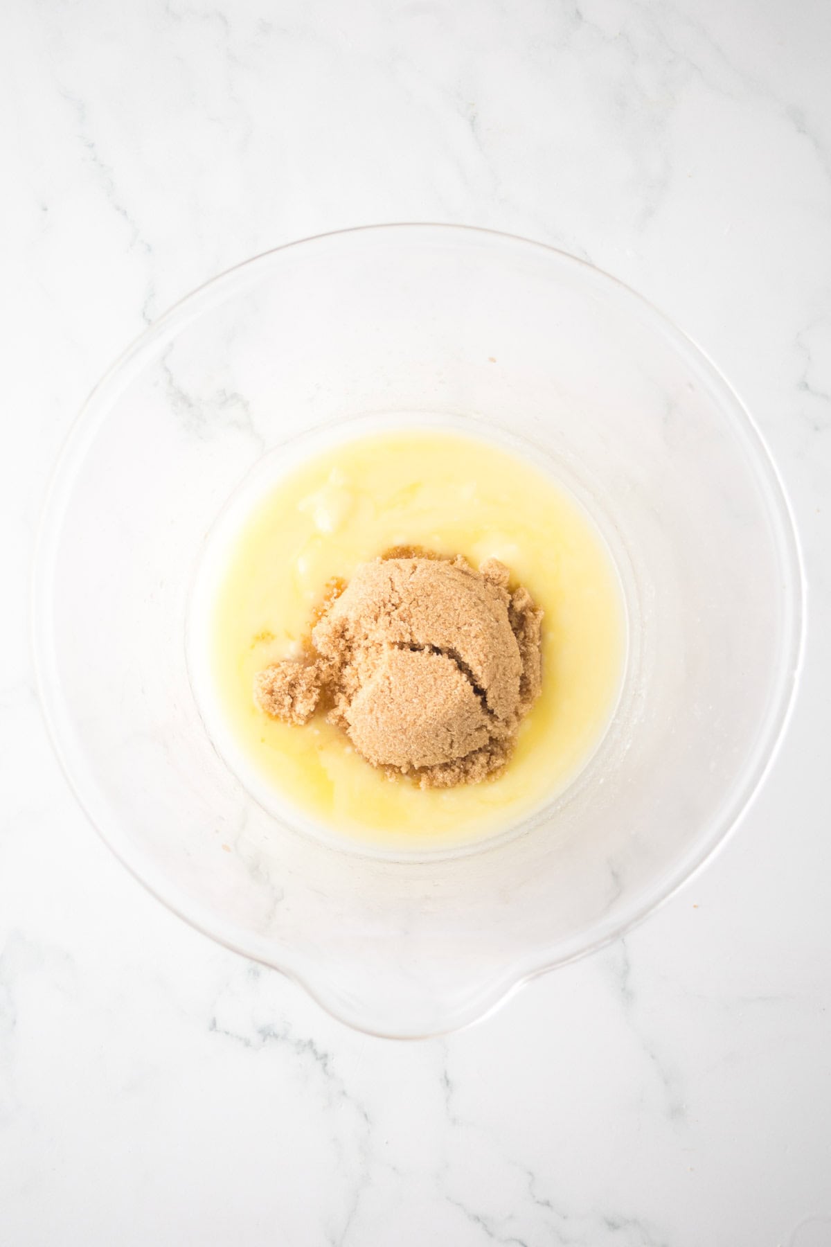 A glass bowl contains a mixture of melted butter and brown sugar on a white marble surface.