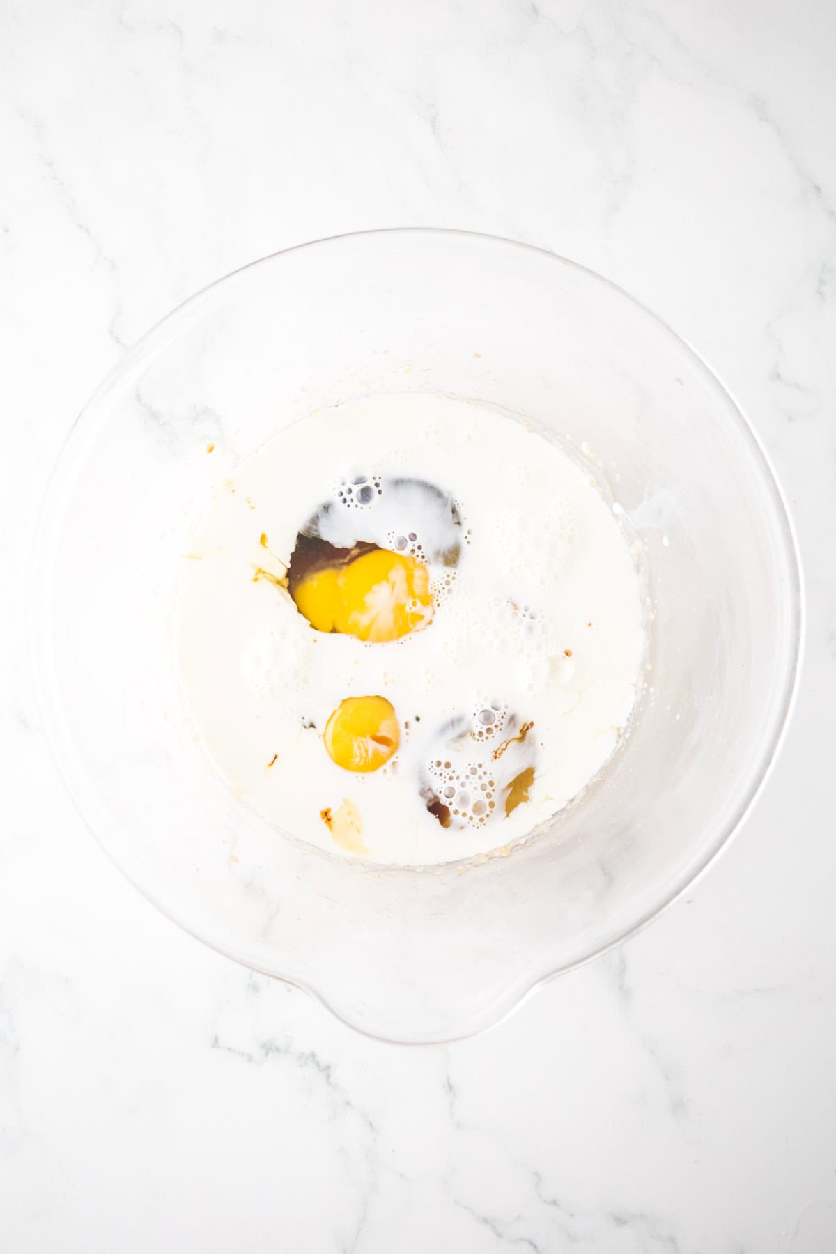A glass bowl contains milk, cracked eggs, and vanilla extract on a white marble surface. The ingredients are ready to be mixed, with the eggs and dark vanilla creating a contrast against the milky liquid.