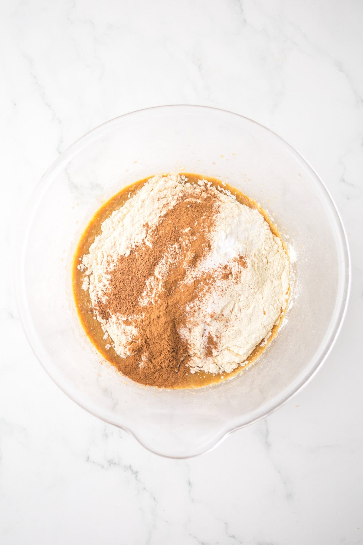 A clear mixing bowl contains a combination of flour, cinnamon, and wet ingredients, ready to be mixed. The bowl is placed on a white marble countertop.