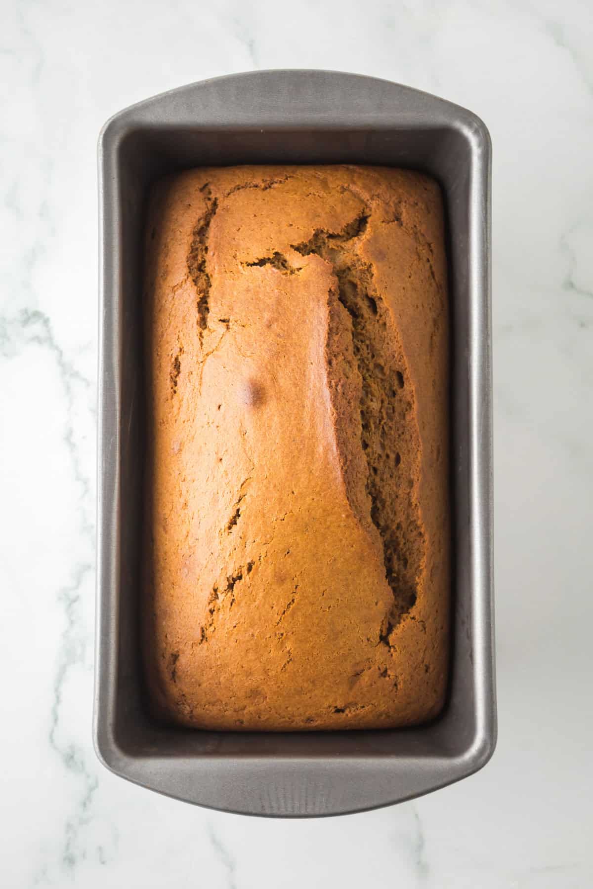 A freshly baked gingerbread loaf in a rectangular metal baking pan on a marble surface. The bread has a golden-brown crust with a prominent crack on the top.