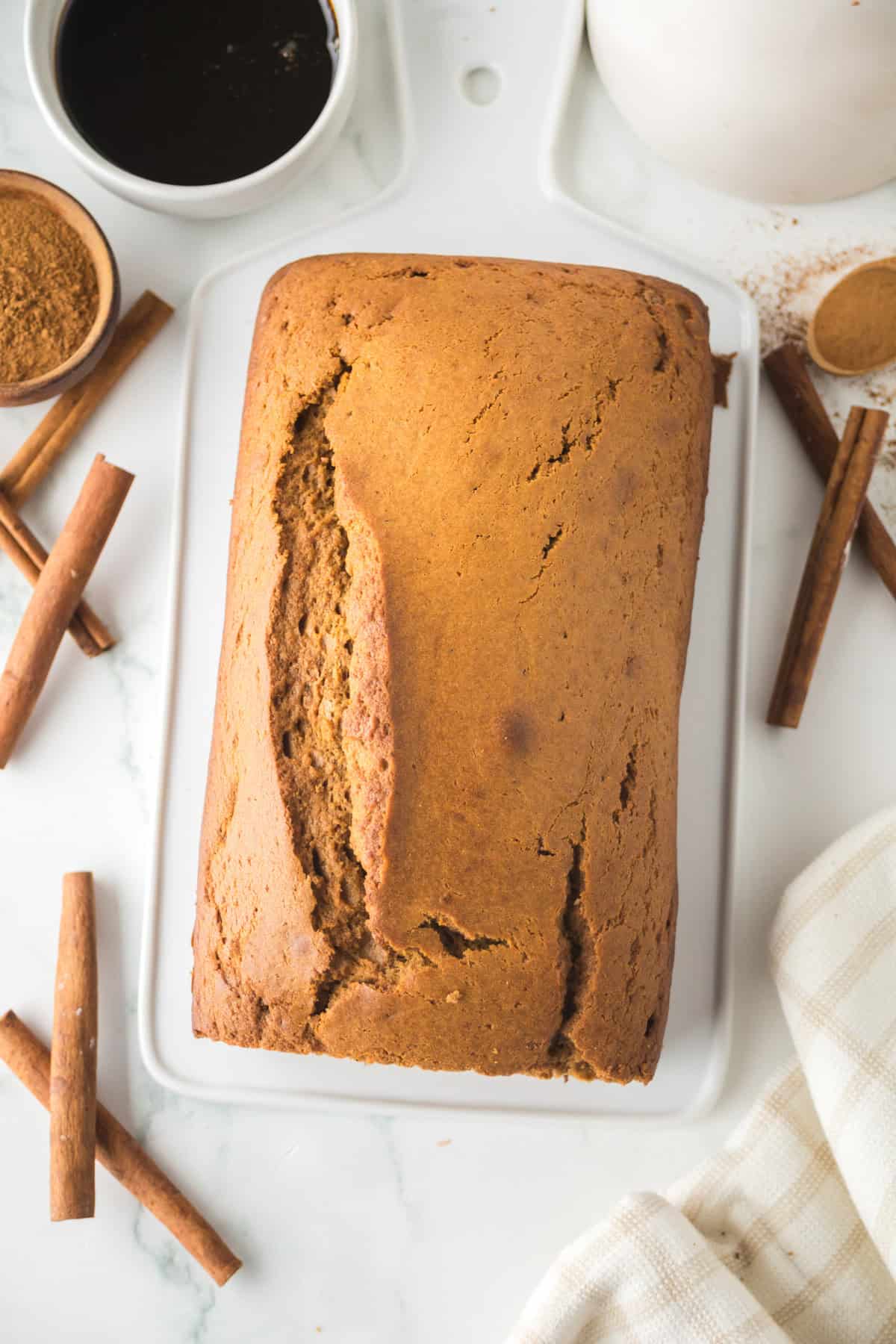 Freshly baked gingerbread loaf rests on a white cutting board, surrounded by cinnamon sticks and a small bowl of cocoa powder. A cup of dark liquid and a white cloth are nearby, set on a white marble surface.