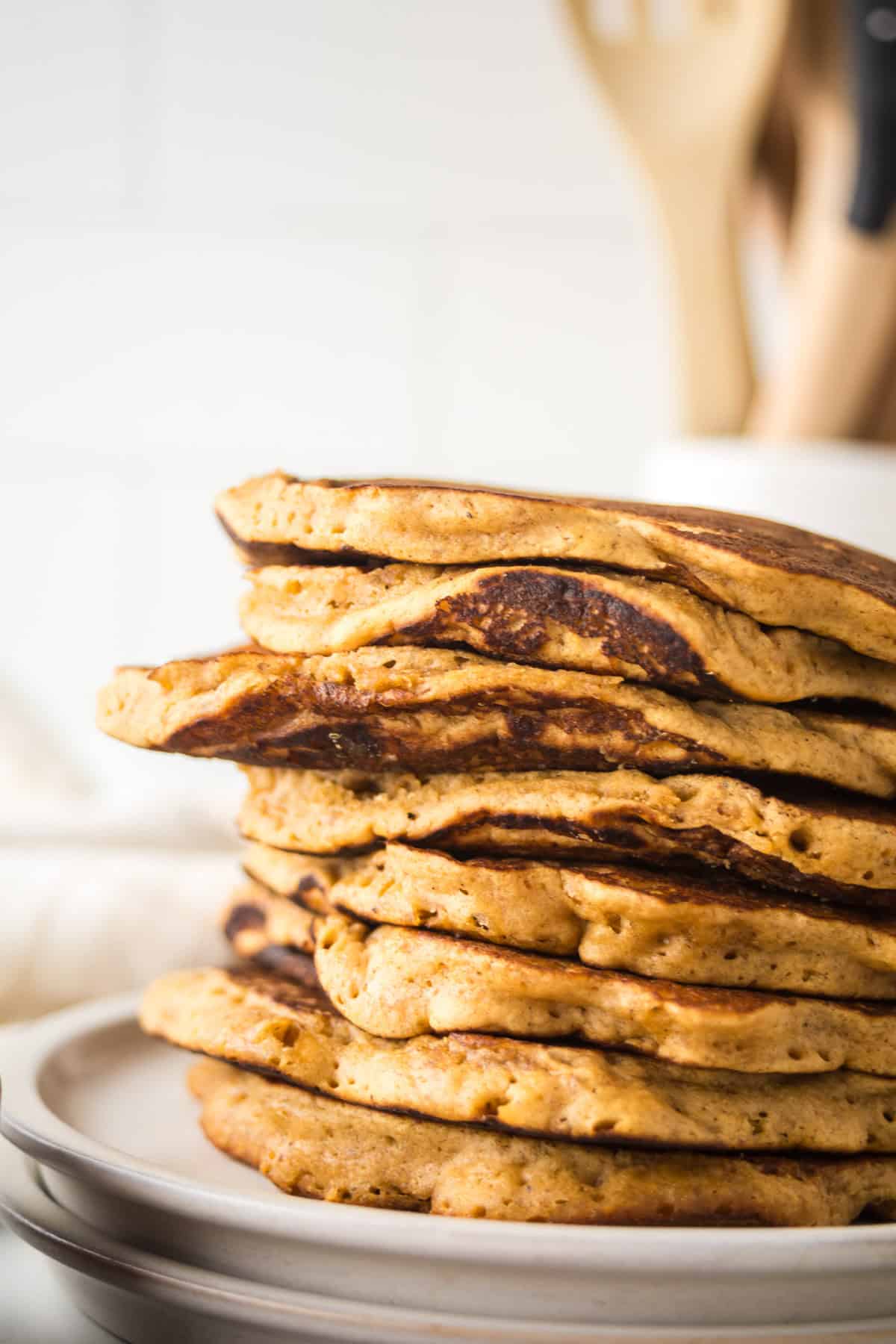 A stack of golden-brown gingerbread pancakes on a white plate, with a blurred background of wooden kitchen utensils in a holder. The pancakes have a fluffy texture and a slightly crisp edge.