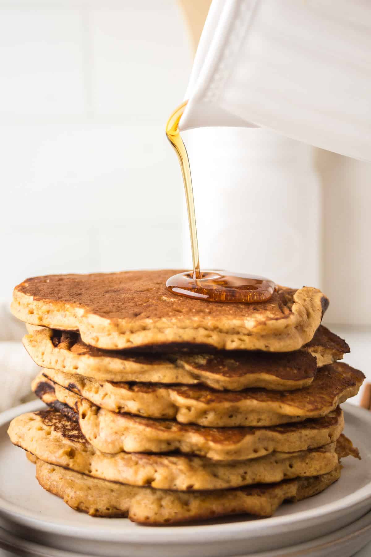 A stack of pancakes is topped with syrup being poured from a white jug. The pancakes are golden-brown and fluffy, sitting on a white plate with a blurred kitchen background.