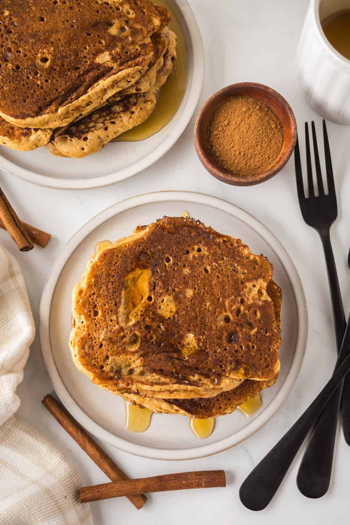 Top view of a plate with stacked cinnamon pancakes drizzled with syrup. Nearby are cinnamon sticks, a bowl of cinnamon powder, a fork and knife, a white cup, and a beige napkin, all on a white marble surface.
