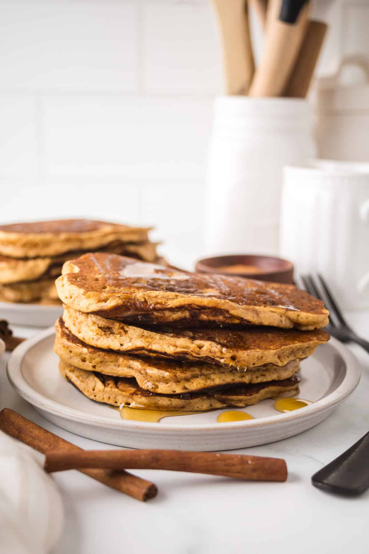 A stack of pancakes drizzled with syrup on a white plate, surrounded by cinnamon sticks. The background includes a blurred white kitchen setting with cooking utensils and containers.
