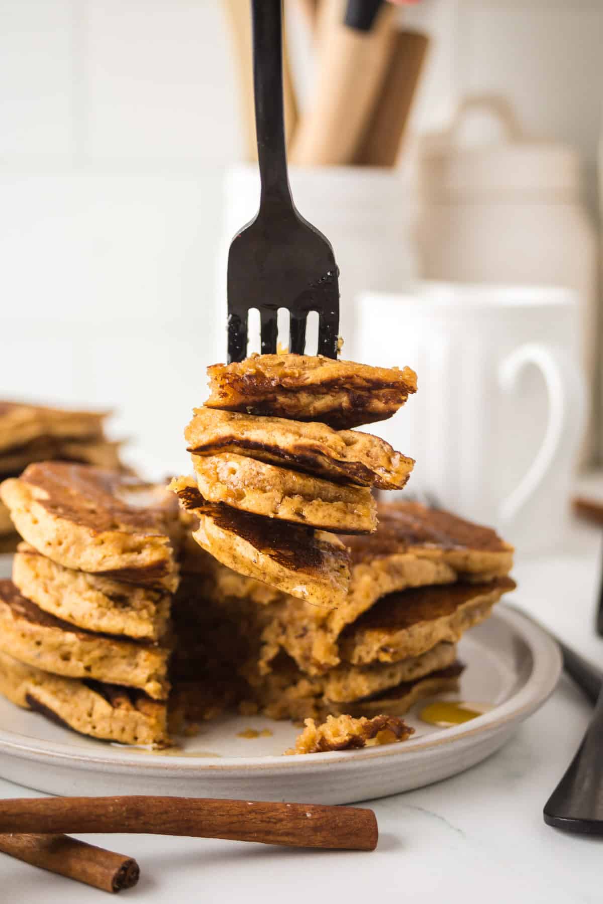 A stack of golden-brown pancakes on a plate is being partially lifted by a black fork. Some pancakes have been cut and syrup is drizzled over them. Cinnamon sticks and kitchen utensils are visible in the blurred background.