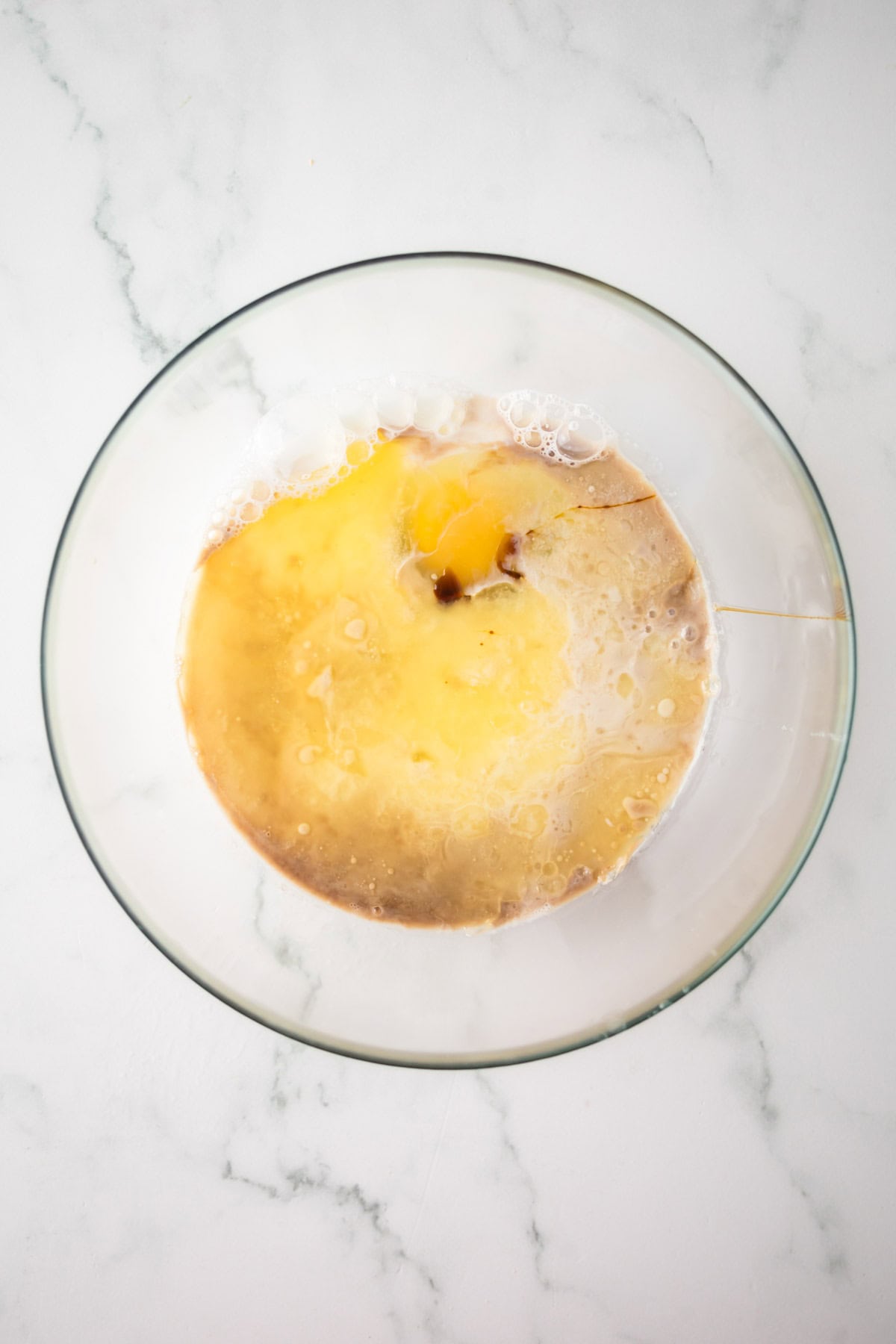 A glass bowl on a marble countertop contains wet ingredients for baking, including cracked eggs, vanilla extract, milk, and melted butter. The mixture is not yet stirred, displaying distinct colors and textures.