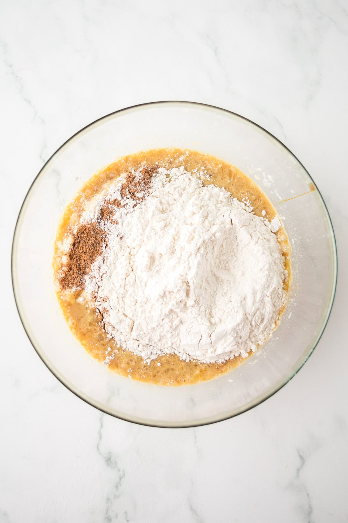 A glass bowl containing a mixture of wet and dry ingredients, including flour, sugar, and spices, on a white marble countertop. The ingredients appear ready to be mixed together.