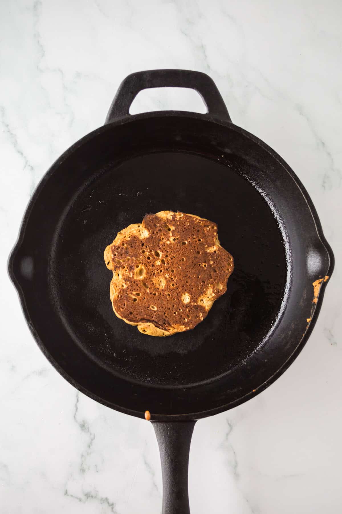 A single pancake cooking in a black cast iron skillet, viewed from above on a white marble surface. The pancake is golden brown with a textured, bubbly surface.