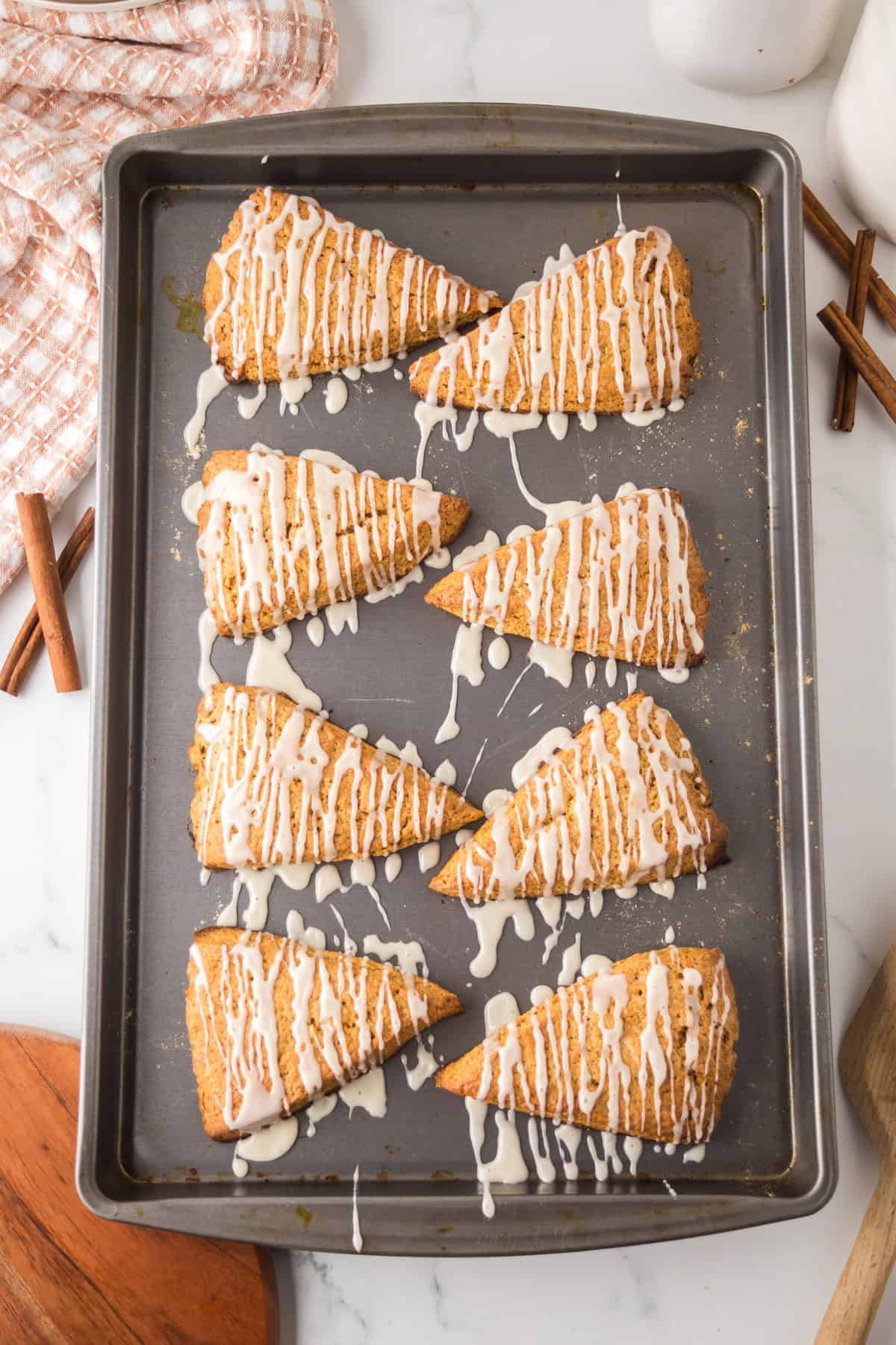 Baked scones arranged on a baking sheet, each topped with a white icing drizzle. Cinnamon sticks are nearby, and a checkered cloth partially shows in the top left corner. The background is marble.