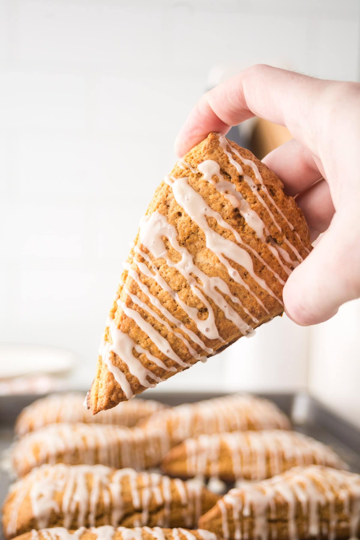 A hand holds a glazed triangular scone, drizzled with white icing. In the background, more scones are on a baking sheet. The scone appears golden-brown, indicating its freshly baked.