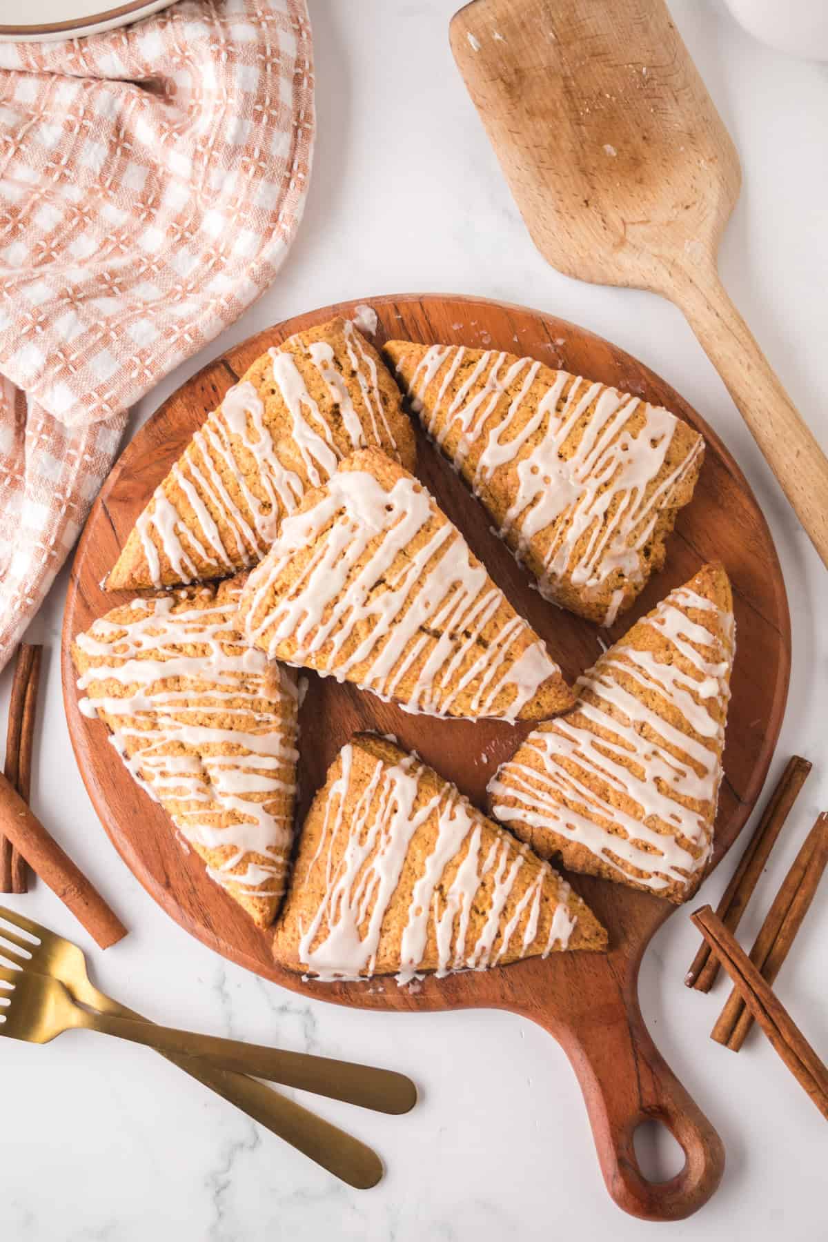 Triangle-shaped gingerbread scones with white icing drizzle sit on a round wooden board. A wooden spatula, checkered cloth, cinnamon sticks, a glass, and gold utensils surround the board on a marble surface.