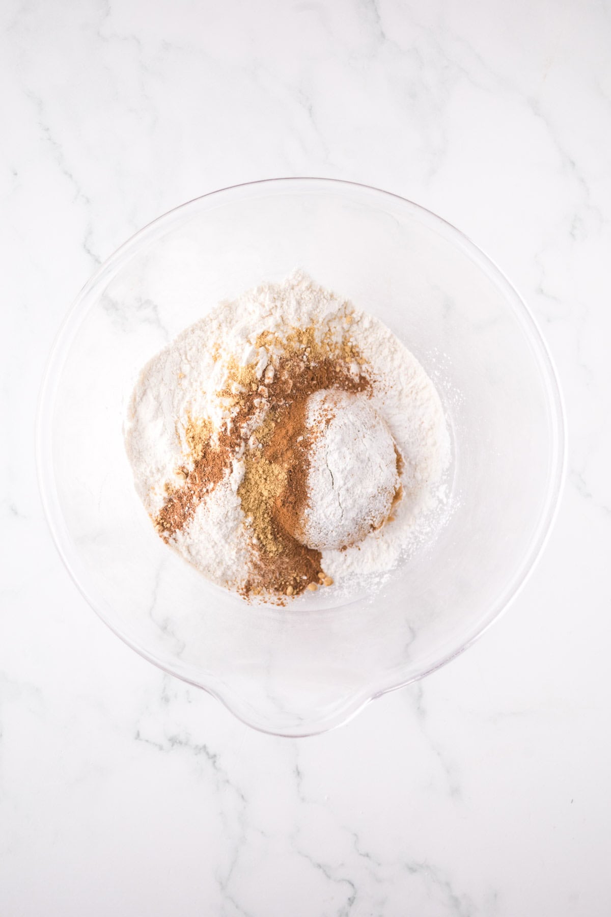 A glass bowl on a marble surface contains various dry ingredients, including flour and brown spices like cinnamon and nutmeg, arranged in small piles.