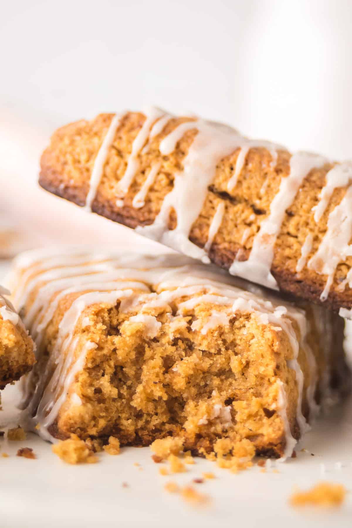 Close-up of pumpkin biscotti drizzled with white icing. One biscotti is leaning on others, revealing a crumbly texture. The image focuses on the contrasting golden brown biscotti and creamy icing.
