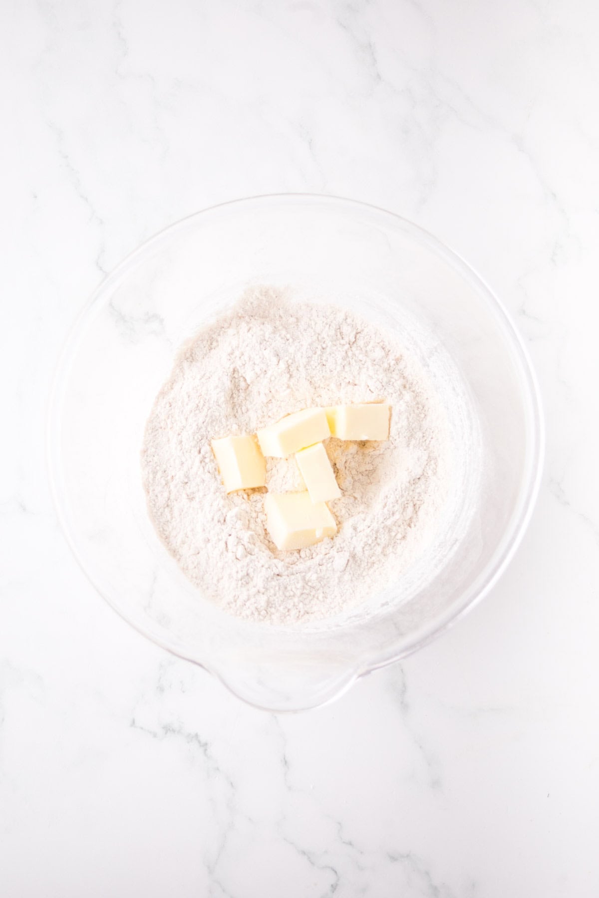 A clear glass bowl contains flour and several chunks of butter on a white marble surface, ready for mixing in baking preparation.