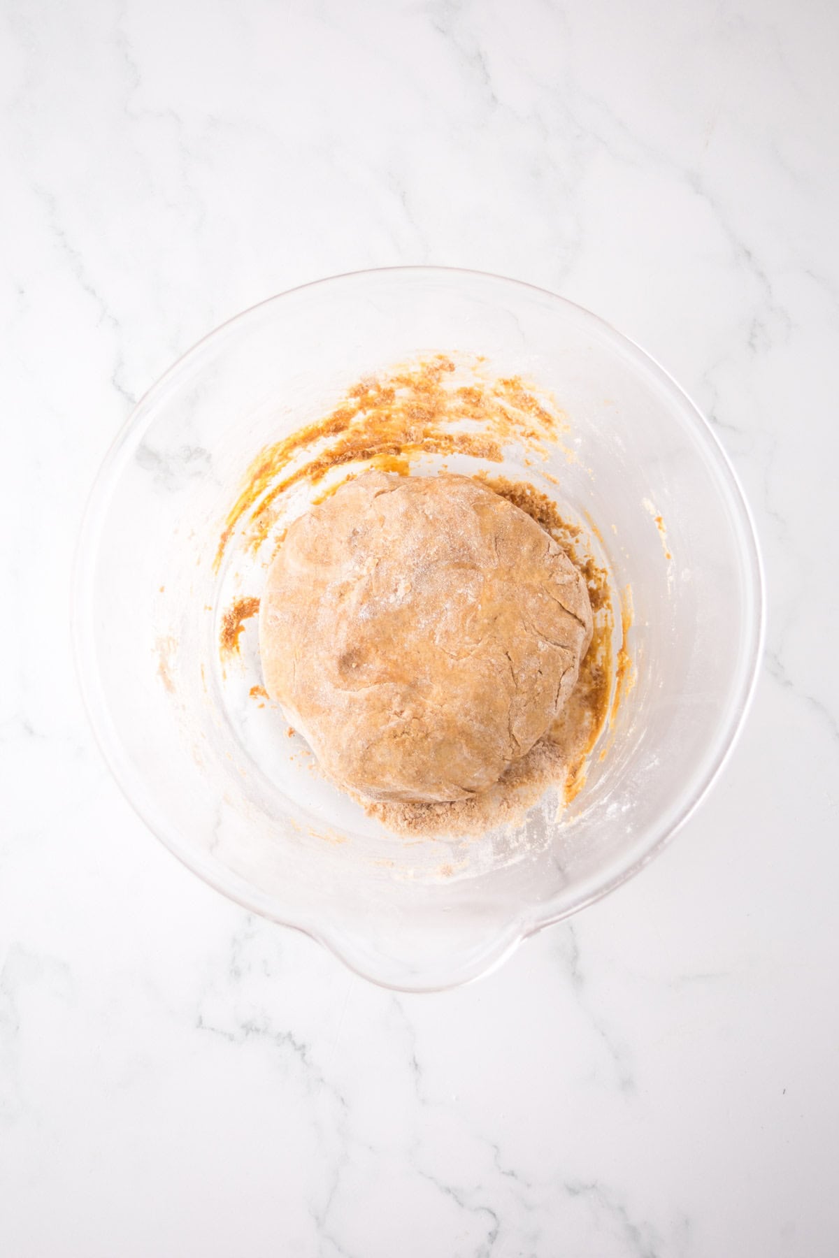 A glass bowl containing a ball of dough is placed on a white marble surface. The dough has a slightly rough texture with a dusting of flour on top. Smudges of dough cling to the side of the bowl.