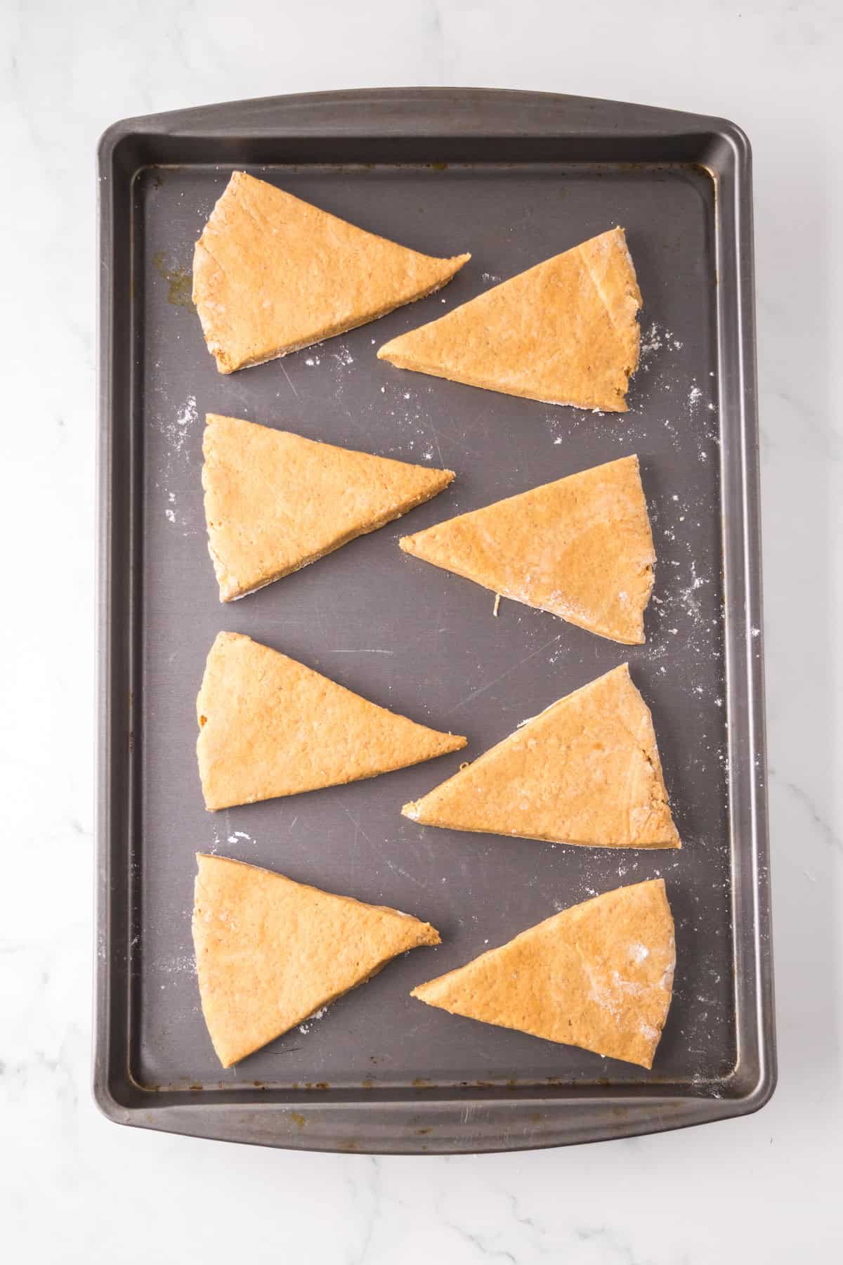 Eight triangular pieces of raw scone dough are evenly spaced on a rectangular baking sheet. The dough is lightly dusted with flour, and the baking sheet rests on a marble surface.