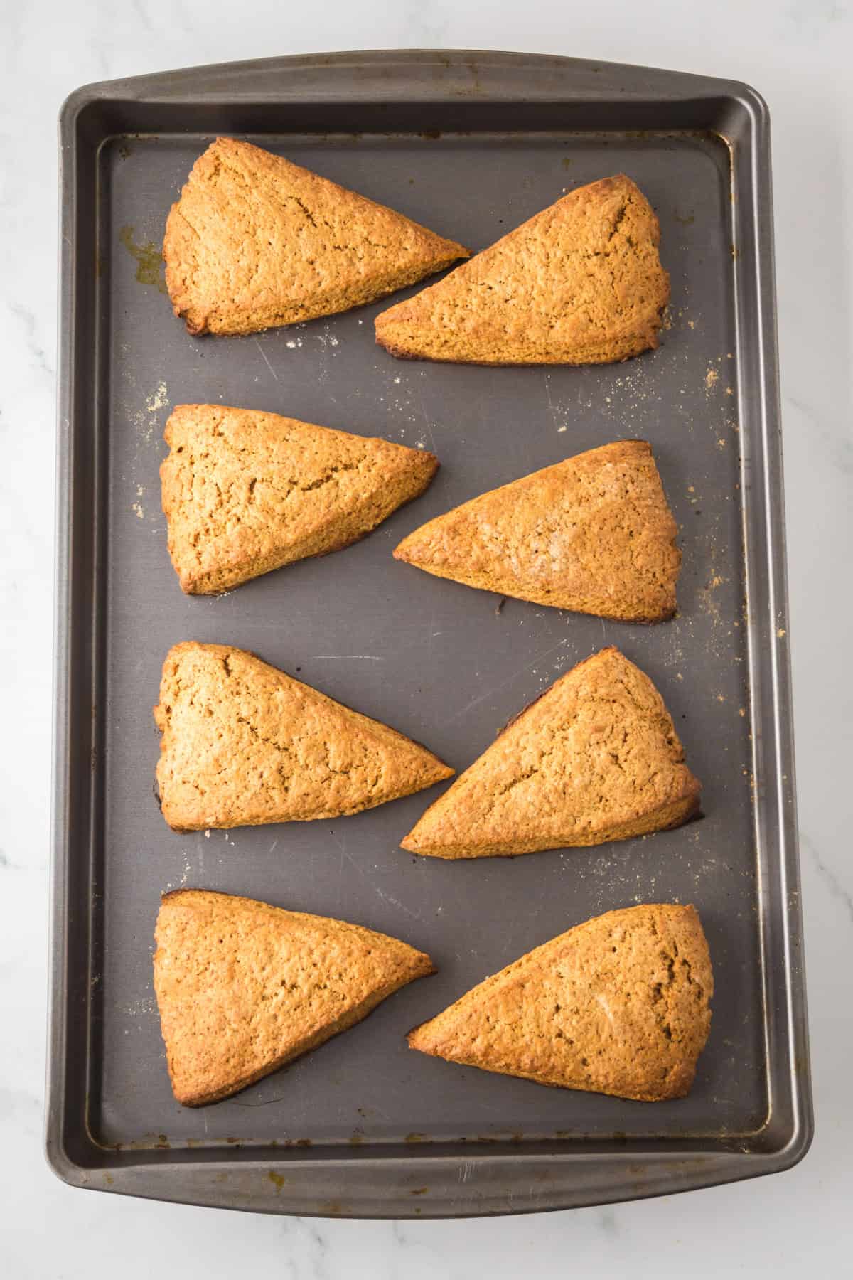Eight triangular, golden-brown scones are neatly arranged on a dark baking sheet. The scones have a crumbly texture with a slightly uneven surface, indicating a crispy exterior. The baking sheet rests on a light marble surface.