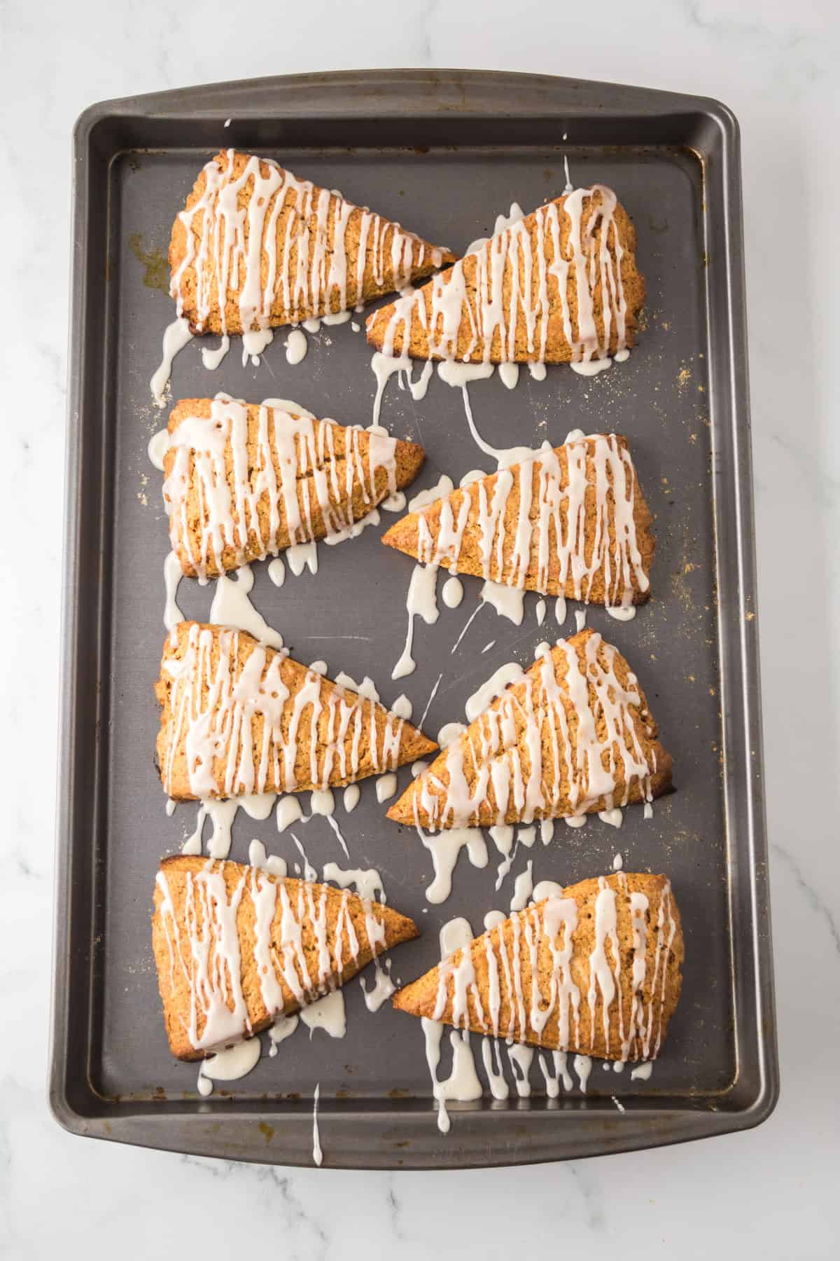 Eight triangular scones drizzled with white icing arranged neatly on a baking tray. The scones have a golden-brown crust and are placed on a marble countertop background.