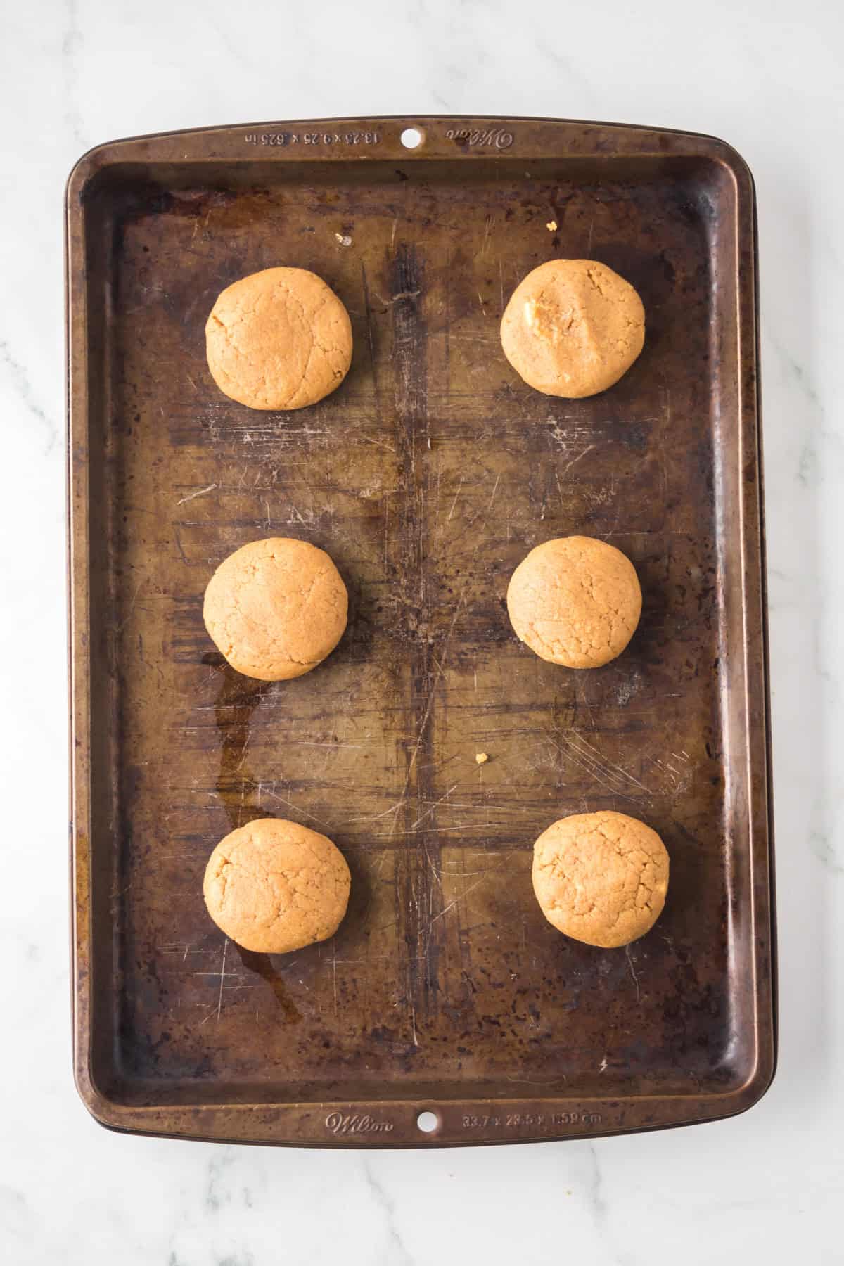 Six round cookies are evenly spaced on a dark, well-used baking sheet. The cookies are golden brown, and the sheet is on a light marble surface.