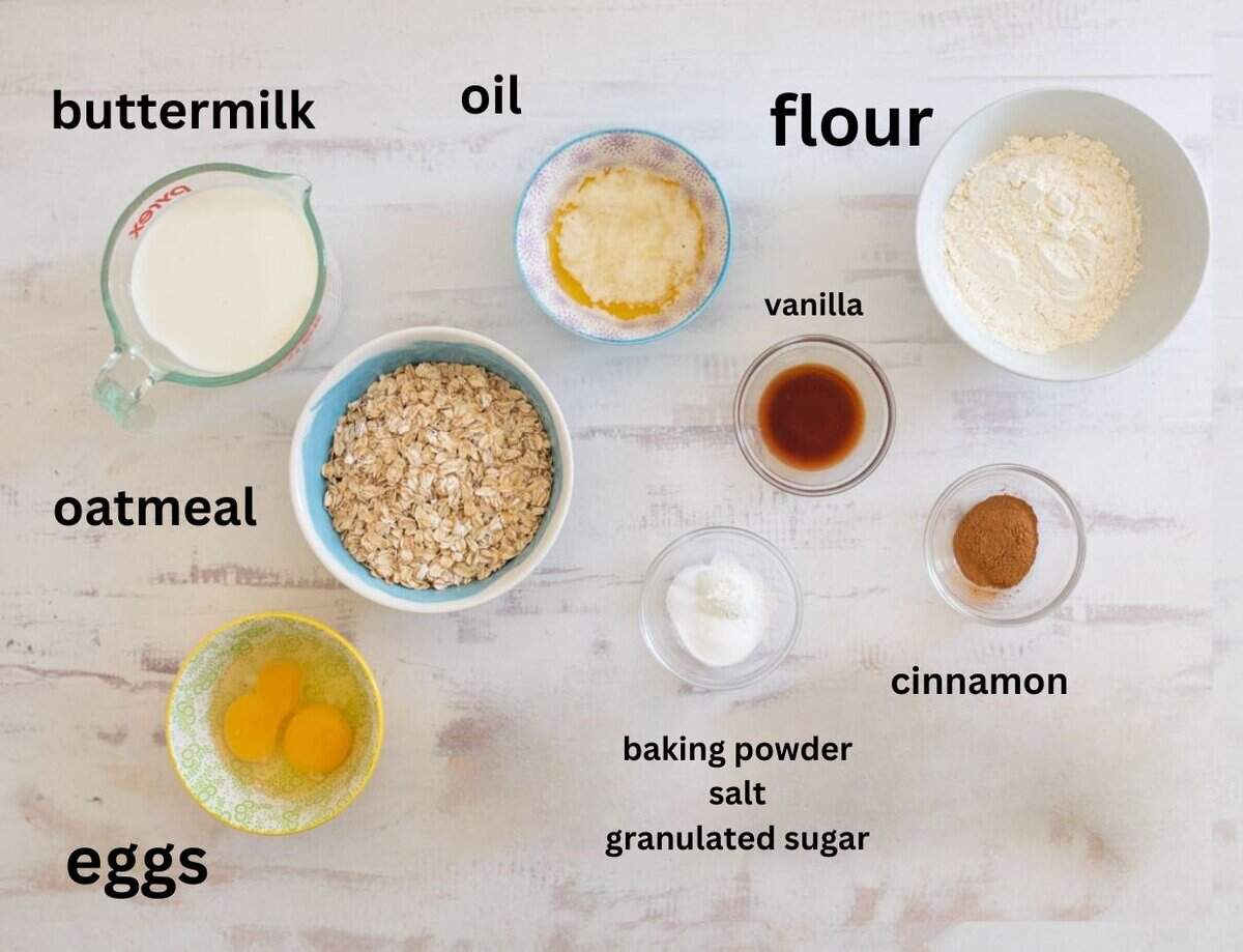 Top view of baking ingredients arranged on a white surface. Items include bowls of buttermilk, oil, flour, oatmeal, eggs, vanilla, cinnamon, baking powder, salt, and granulated sugar. Each item is labeled with text.