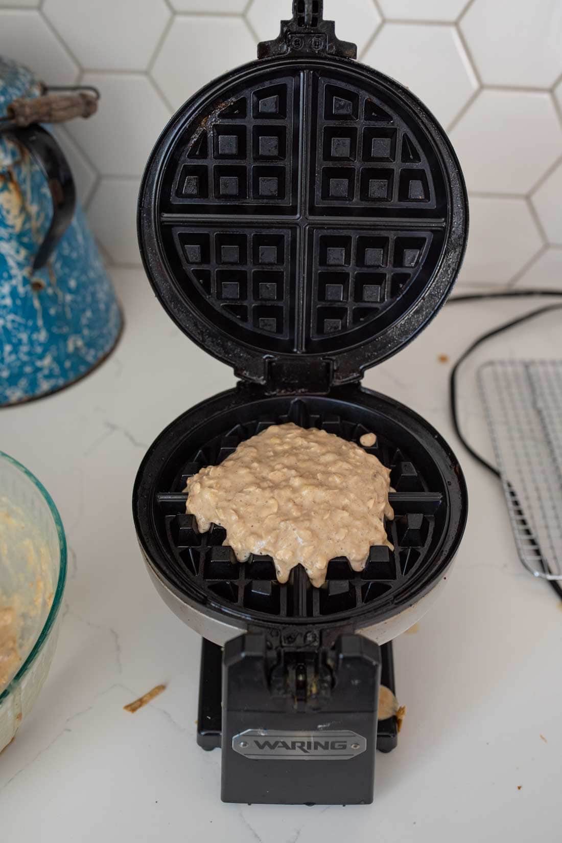 Electric waffle maker with waffle batter spread on the non-stick plates, ready to cook. A vintage blue kettle is seen on the left, and a cooling rack is in the background on a countertop with white hexagonal tiles.