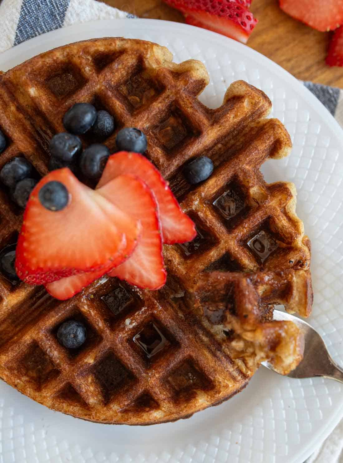 A close-up of a waffle on a white plate, topped with sliced strawberries and blueberries. A fork with a waffle piece is resting on the side. A few more strawberry slices are in the background.