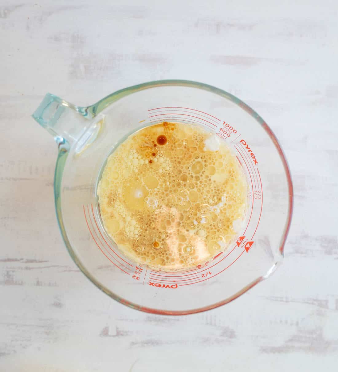 A glass measuring cup filled with a frothy, golden-brown liquid mixture sits on a white surface. Measurements are marked in red along the side of the cup.