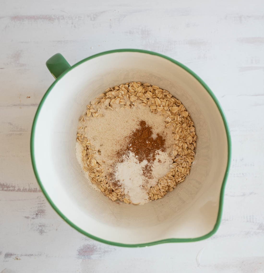 A mixing bowl with green edges containing dry ingredients: oats, sugar, and a dusting of cinnamon or other spices, on a light-colored surface.