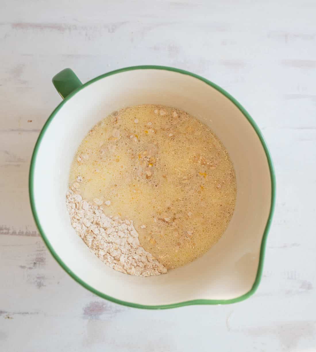 A mixing bowl with green edges containing a blend of dry oats and a wet mixture, partially mixed, on a light wooden surface.