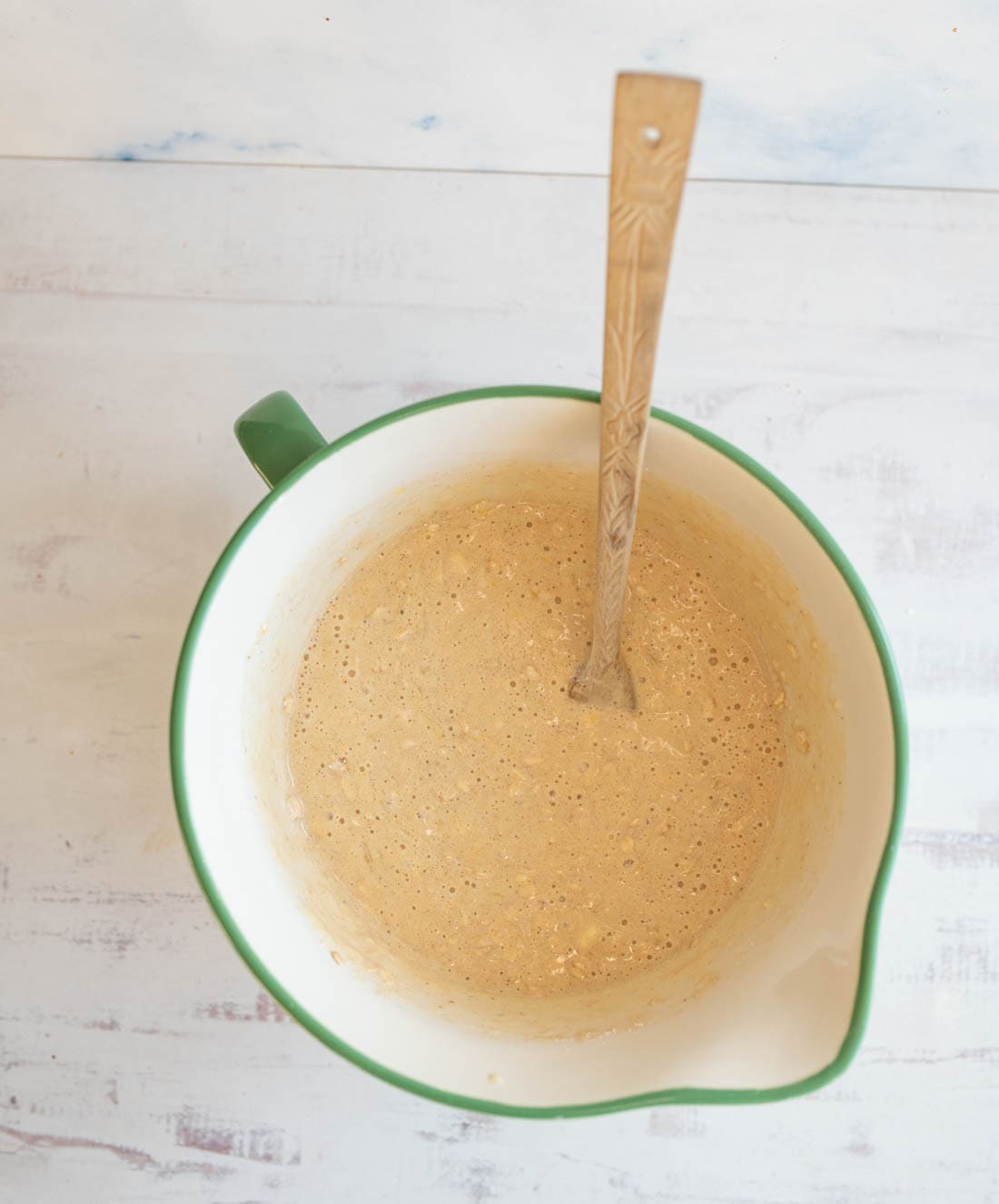 A green-rimmed mixing bowl filled with a light brown batter sits on a wooden surface. A metal spoon is in the batter, ready for stirring or serving. The surface has a light, weathered appearance.
